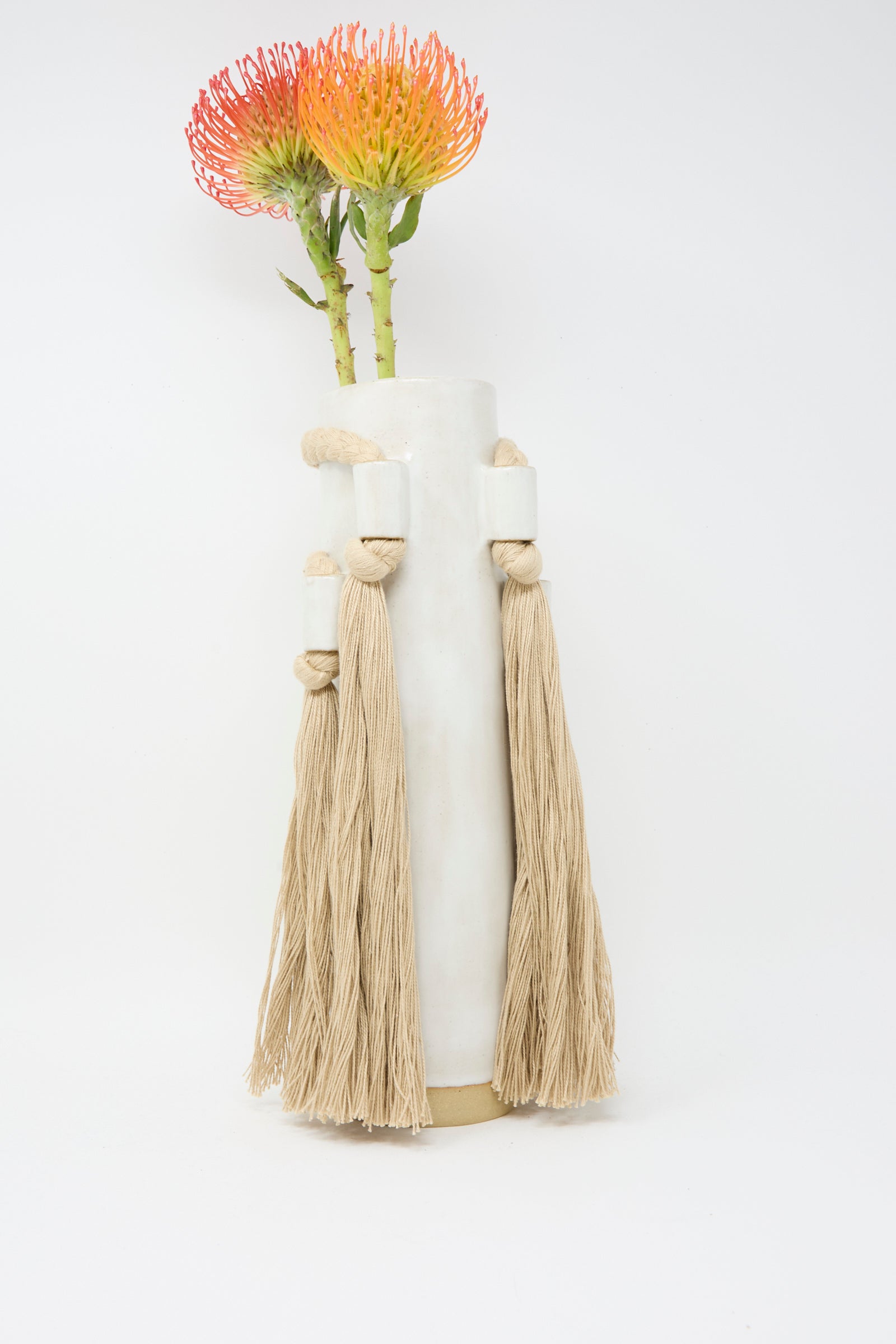 Karen Tinney's Vase #735 in White, a handmade stoneware piece with rope and tassel accents, holds two orange flowers against a plain white background.