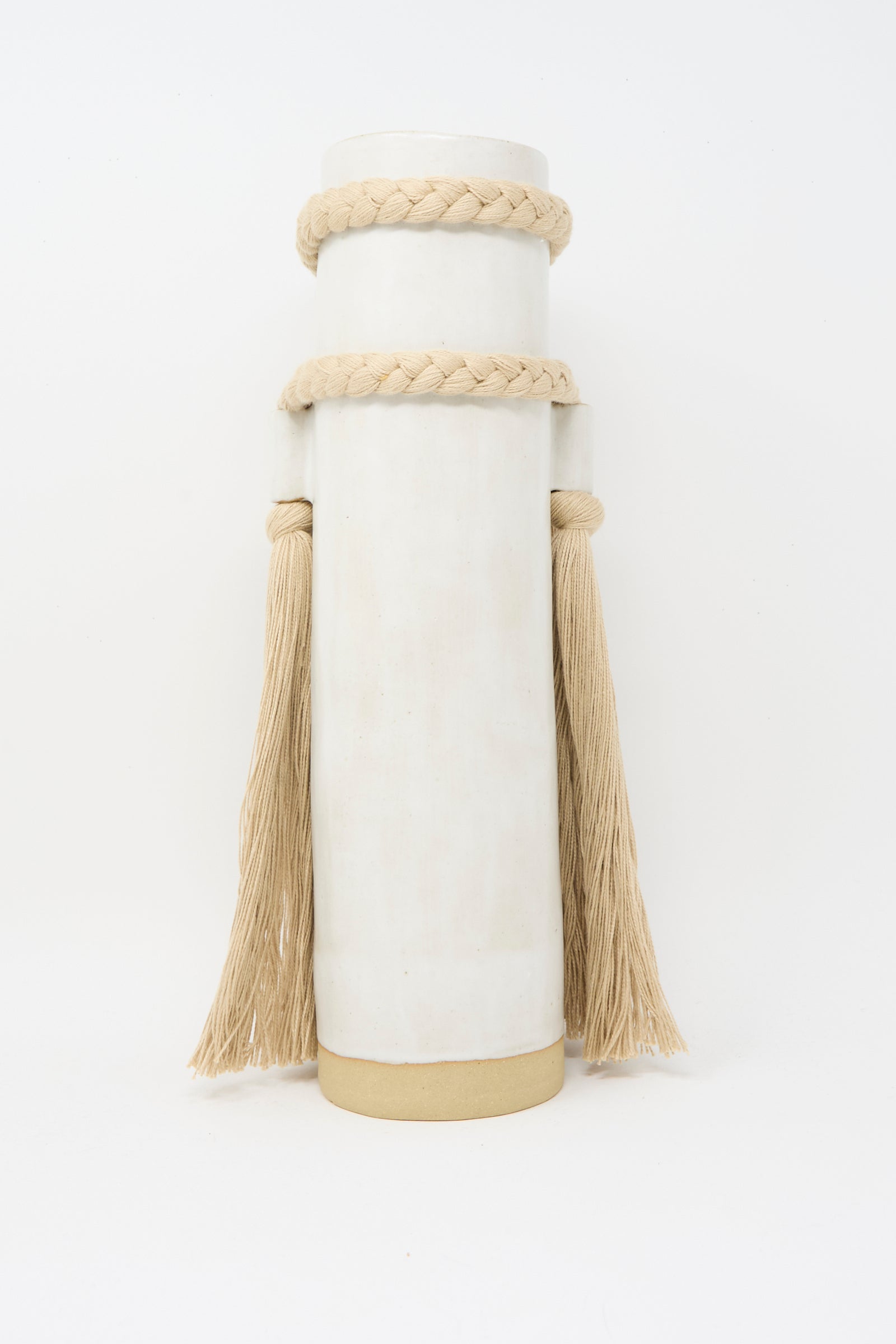 The Karen Tinney Vase #735 in White is a tall, cylindrical, handmade Vermont piece featuring a white glaze, beige base, and braided rope with long tassels draped from each side.