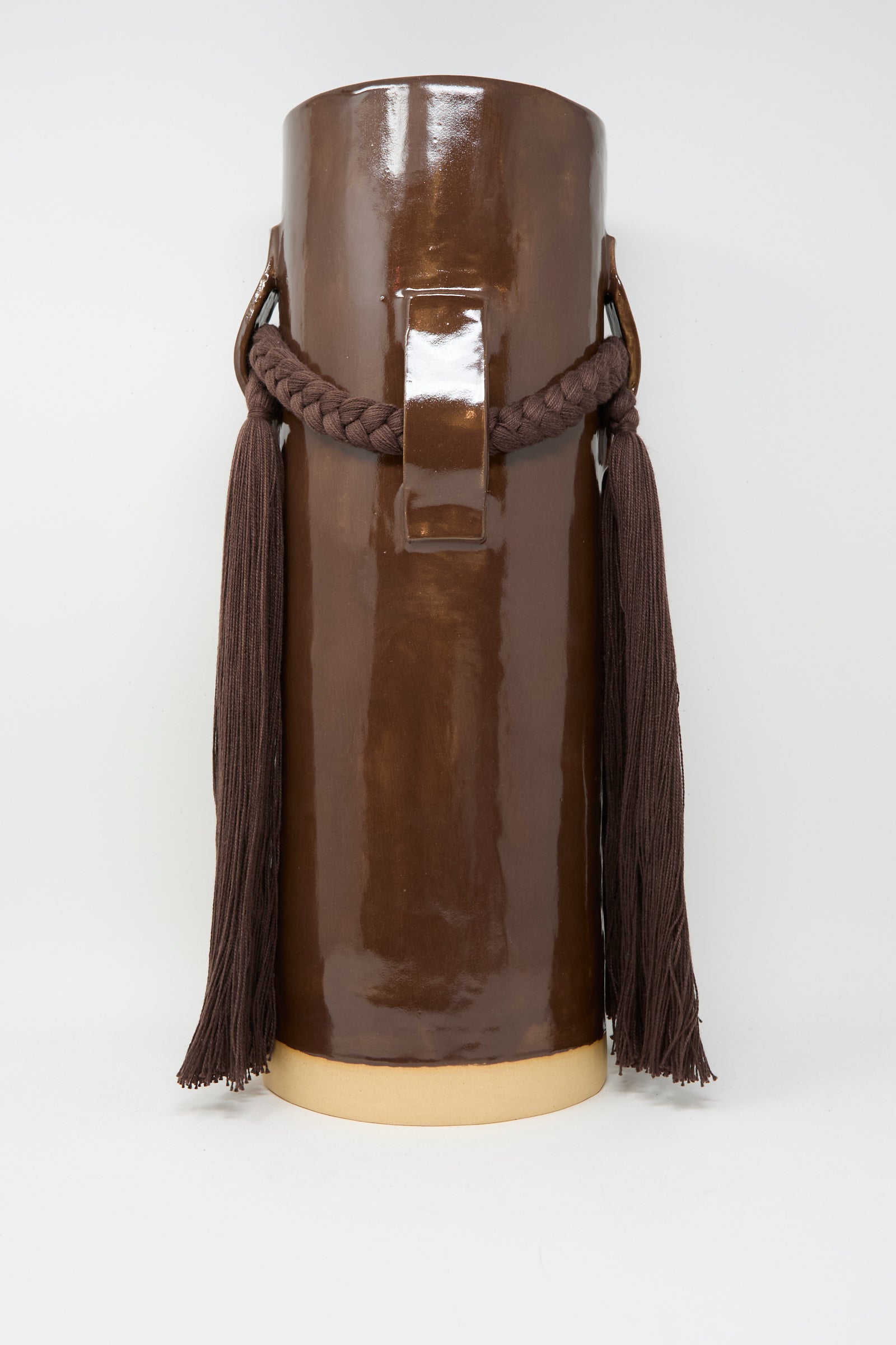 The Karen Tinney Vase #800 in Brown is a tall, cylindrical stoneware vase with a glossy finish, handmade in Vermont. It features braided rope handles and long tassels on each side.