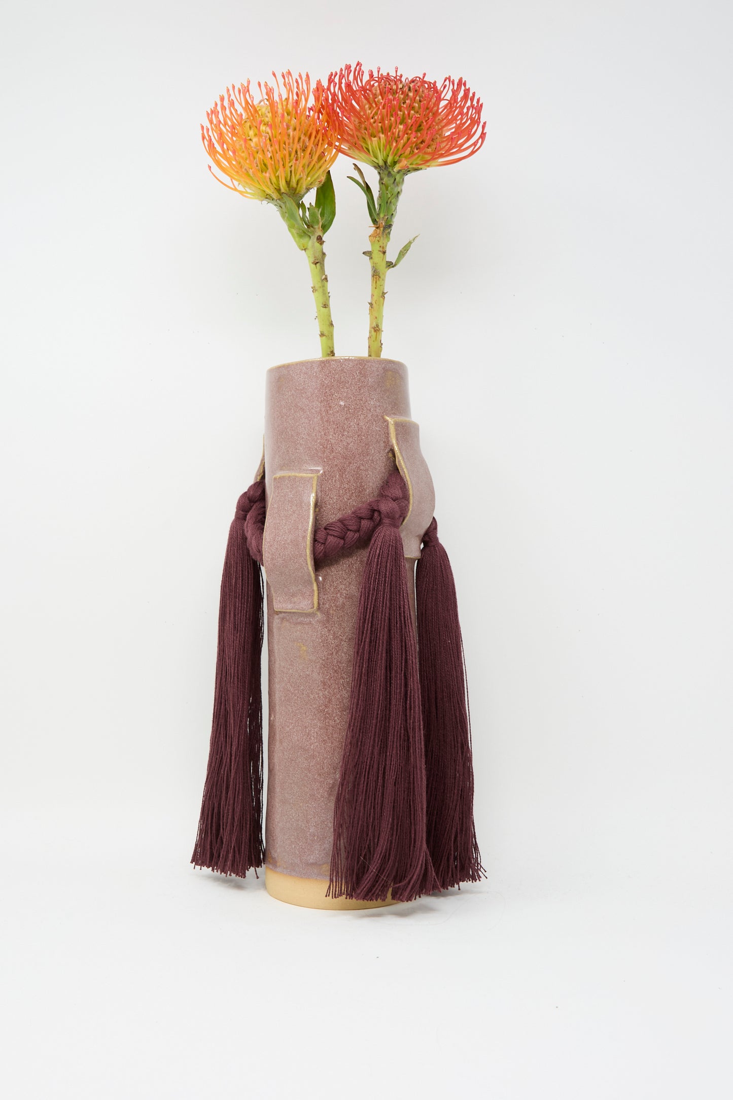 Karen Tinney's Vase #800 in Burgundy, a tall handmade pinkish vase with a braided tasseled cord, holds two orange protea flowers against a plain white background.
