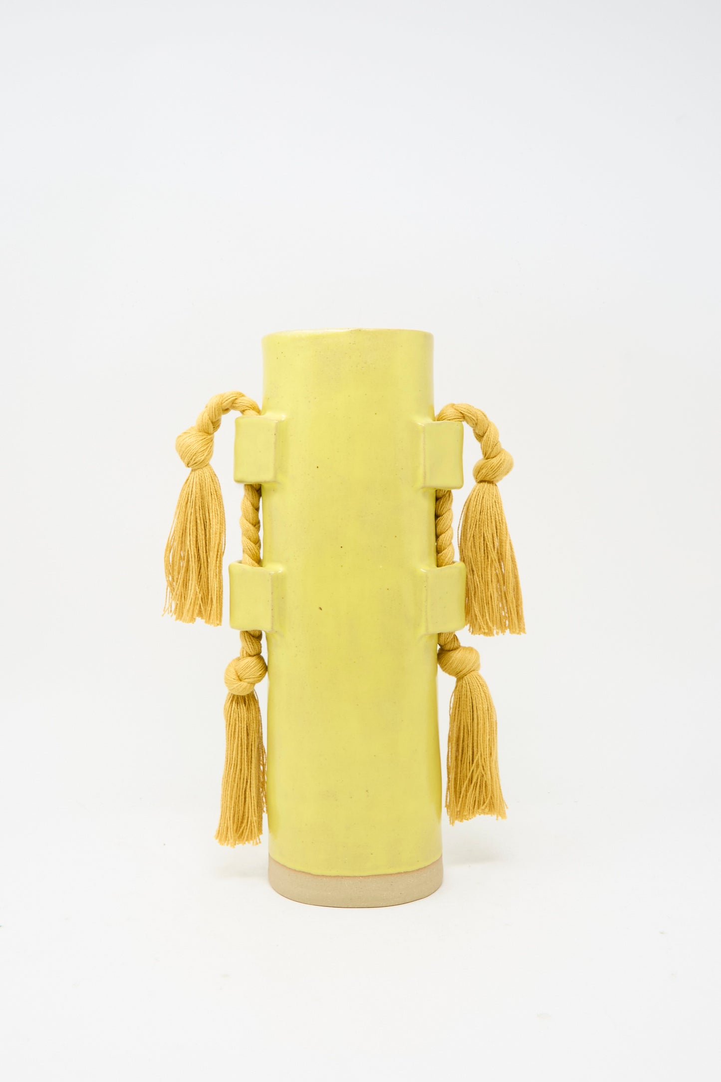 Vase #504 in Marigold by Karen Tinney is a tall, cylindrical vessel featuring four thick rope handles, each accented with a large yellow tassel. Set against a plain white background, this handmade piece adds artisan charm to any space.