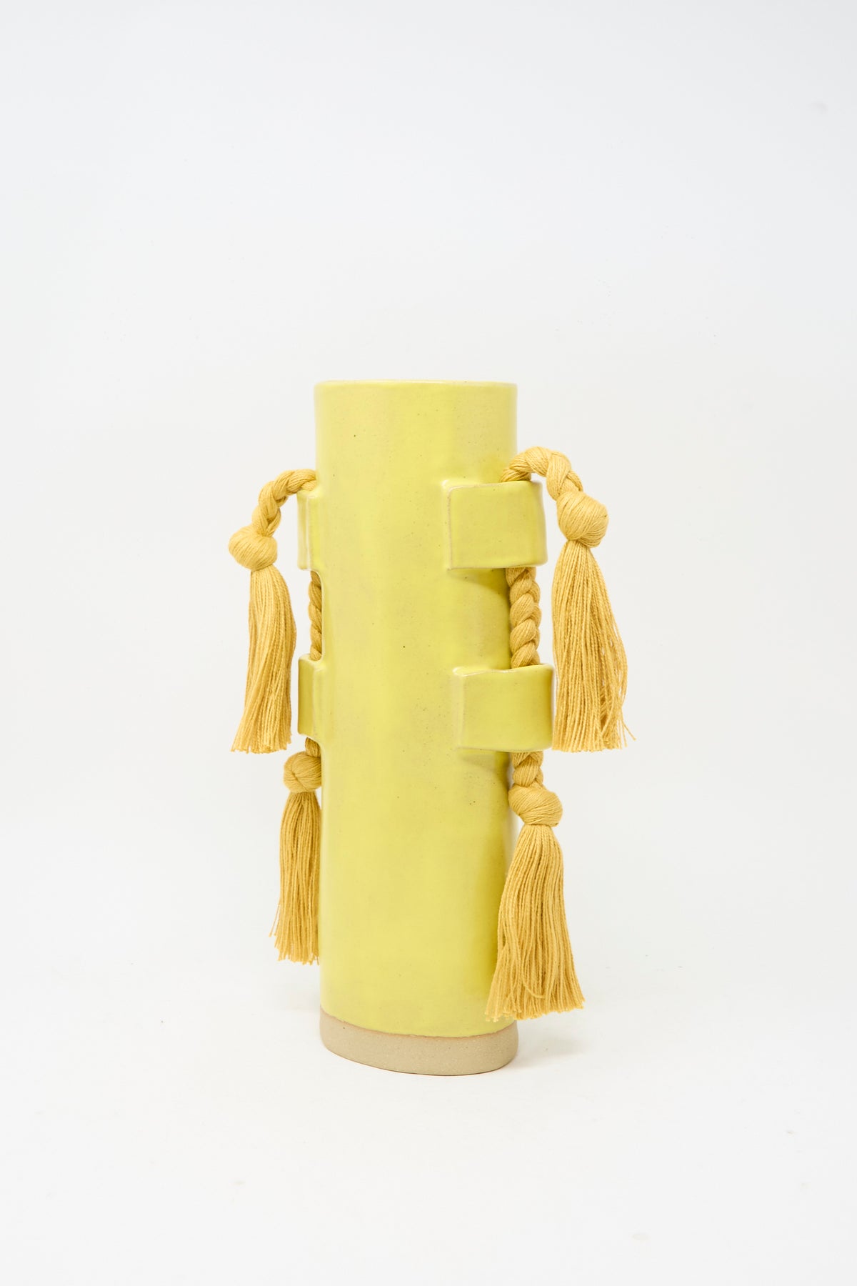Vase #504 in Marigold by Karen Tinney—a tall, cylindrical vase featuring rectangular handles and adorned with thick yellow braided cords and tassels on each side—stands against a plain white background.