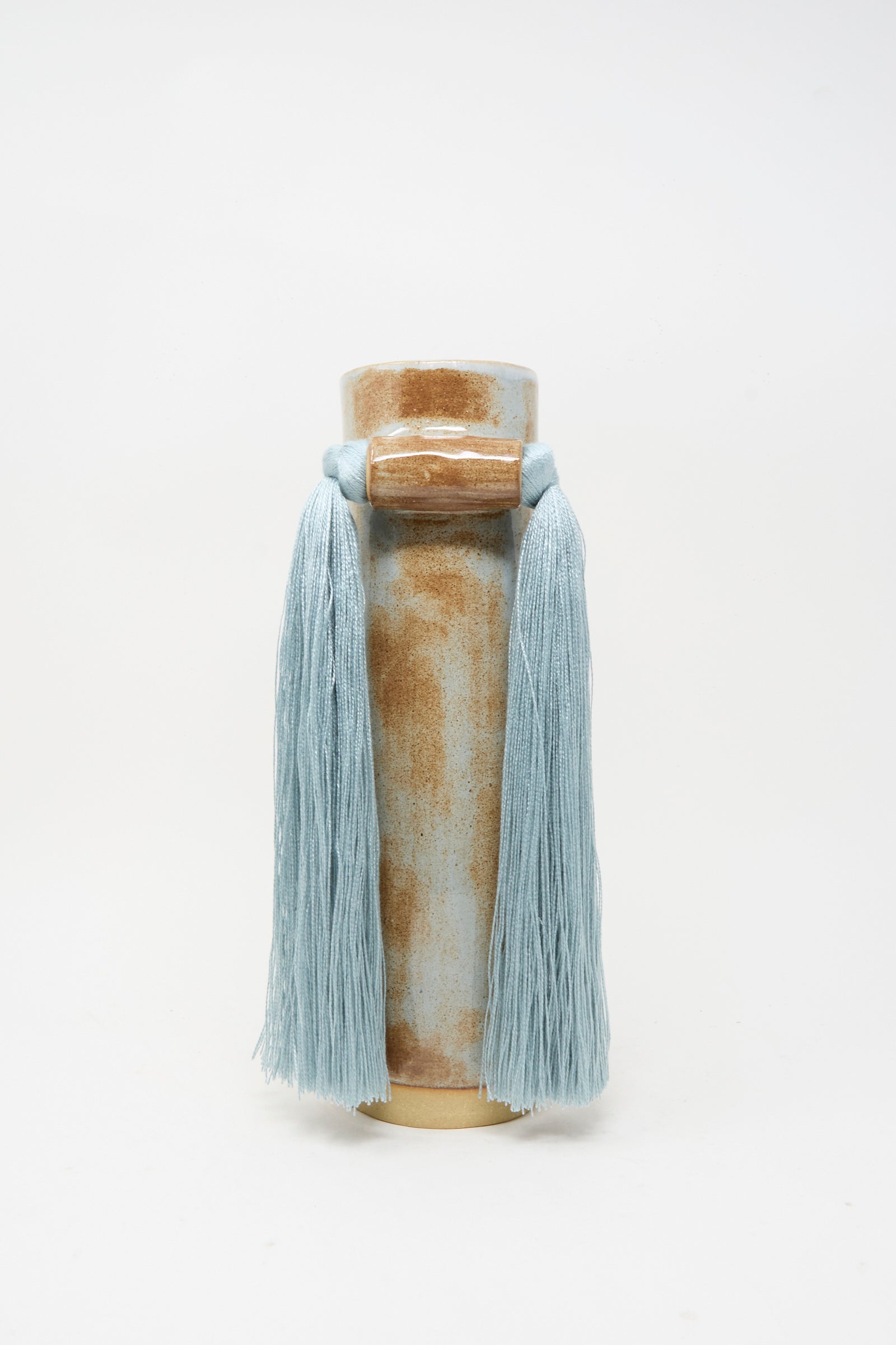 Vase #531 in Blue by Karen Tinney is a tall, handmade stoneware vase with a cylindrical shape, finished in a brown and blue glaze and adorned with long, light blue tassels draped from a central knot near the top.