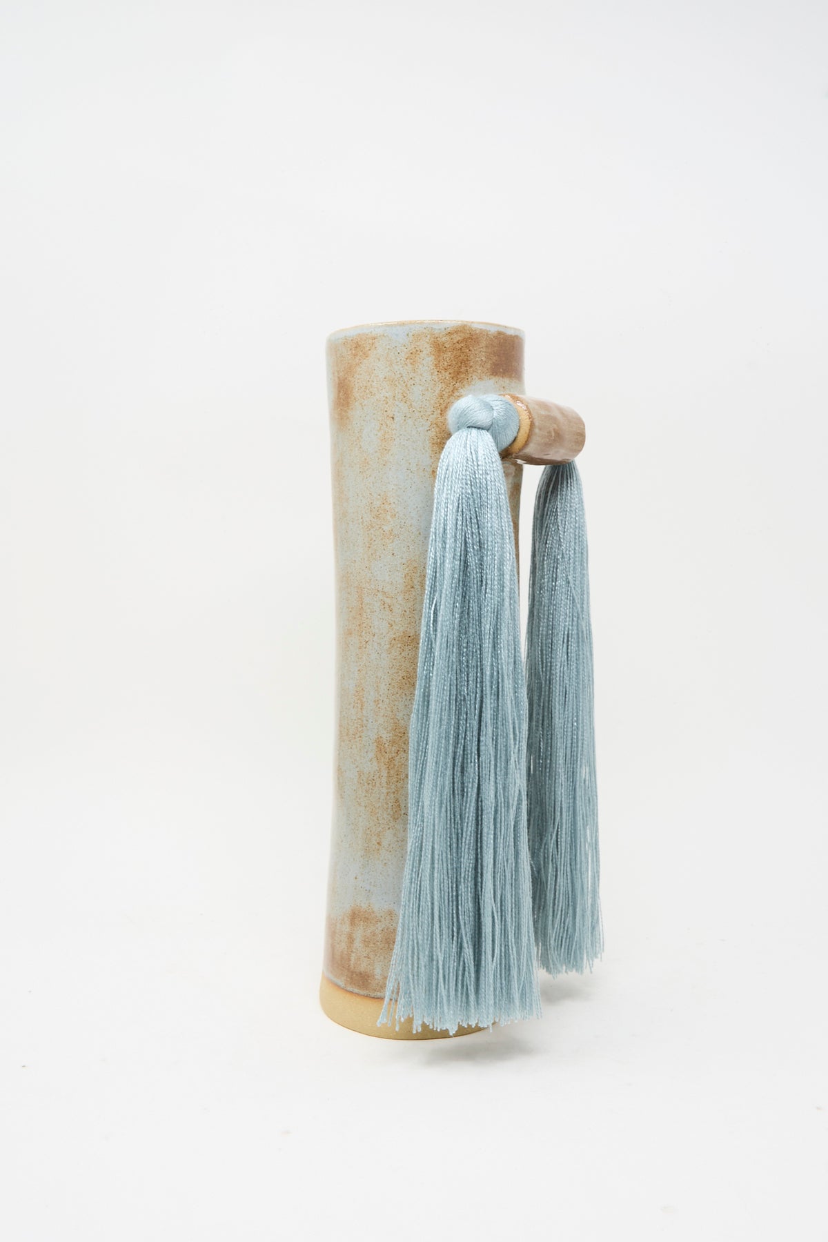 Vase #531 in Blue by Karen Tinney is a tall ceramic vase with a beige and blue mottled finish, reminiscent of handmade stoneware, and features a wooden handle adorned with two long, light blue tassels.