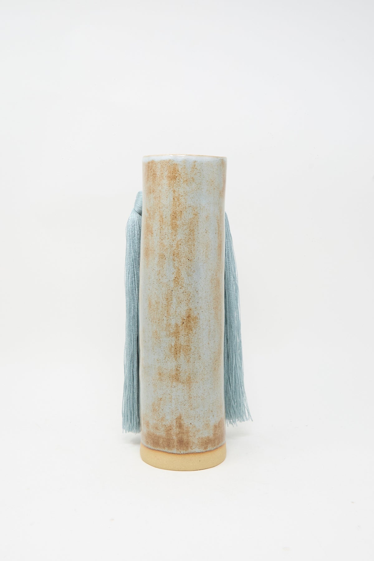 Vase #531 in Blue by Karen Tinney is a tall, cylindrical handmade stoneware vase with a distressed beige and light blue finish. It features long blue tasseled cords hanging from the sides and is set against a plain white background.