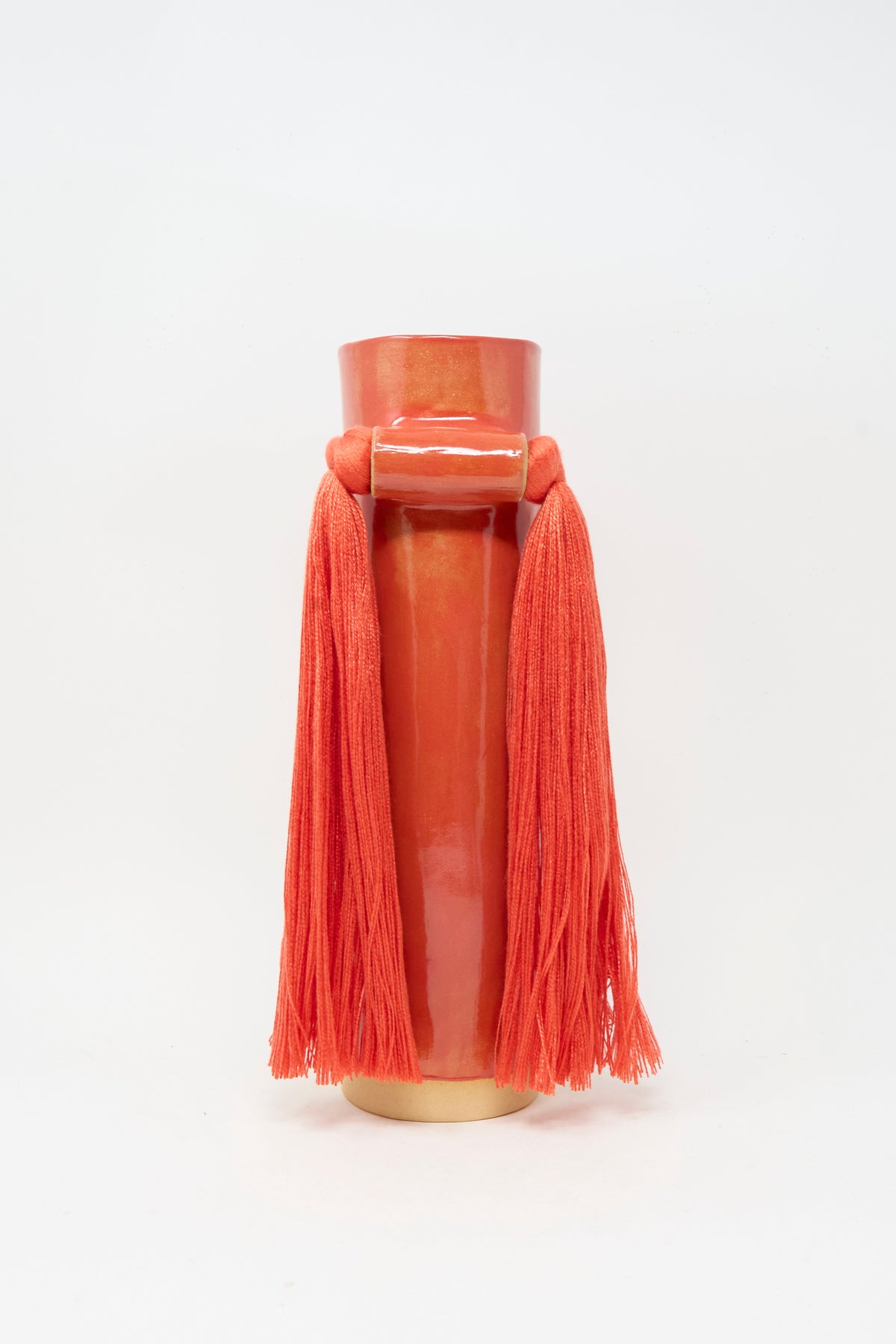Vase #531 in Red by Karen Tinney is a handmade stoneware vase, expertly crafted in the spirit of Vermont pottery. This tall, glossy red ceramic piece features two long tassels hanging from a horizontal bar near the top and is set against a white background.