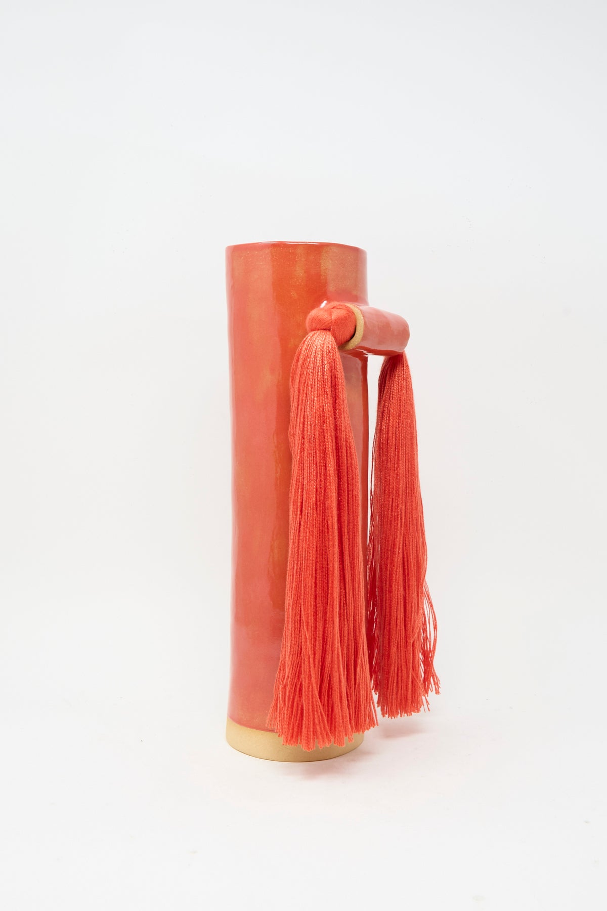 Vase #531 in Red by Karen Tinney is a tall, cylindrical, glossy coral handmade stoneware vase featuring a side handle and two long coral tassels, displayed against a plain white background.