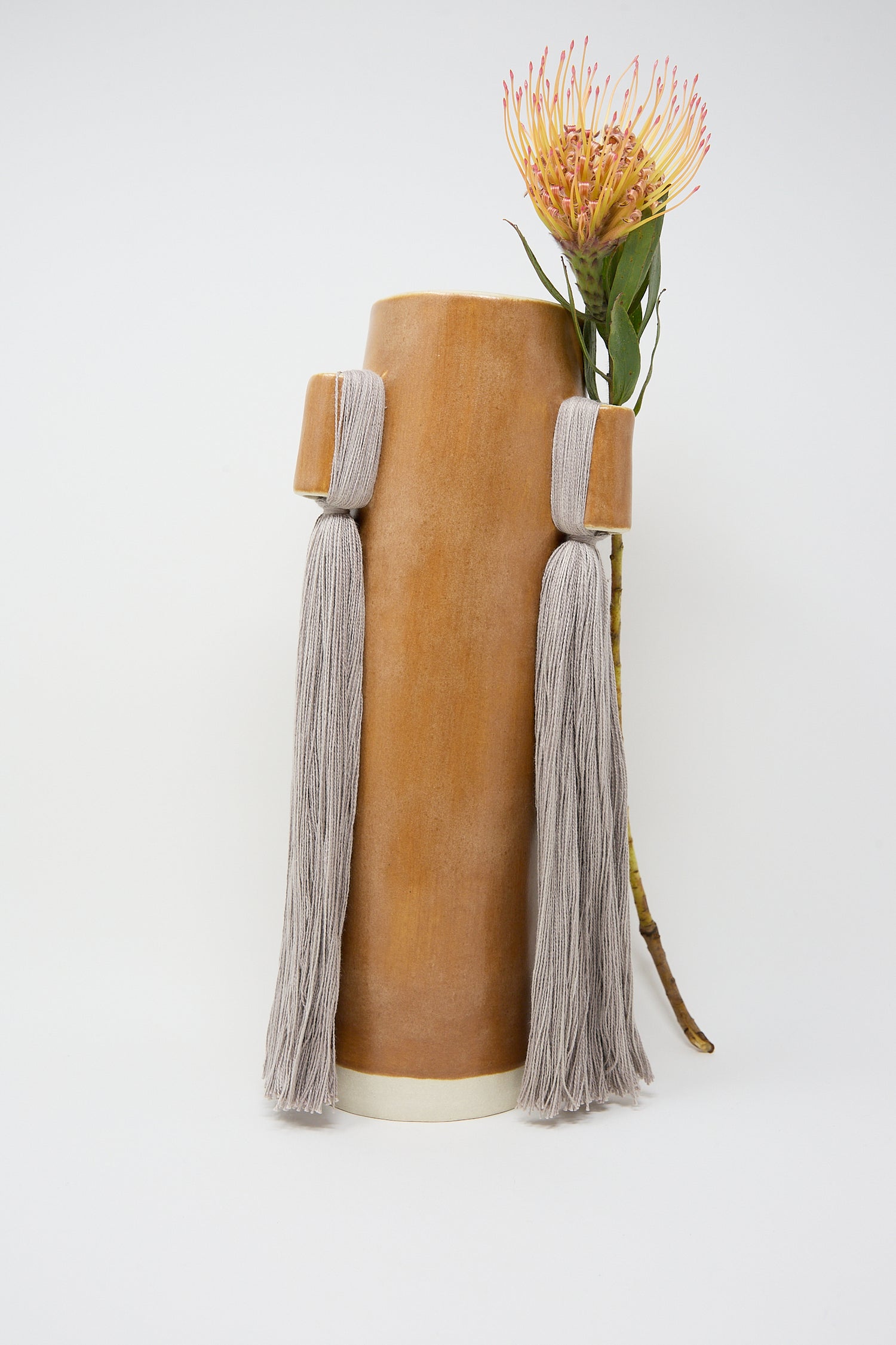 A cylindrical leather Vase #607 in Brown with tassels displaying a single protea flower is handcrafted by Karen Tinney.