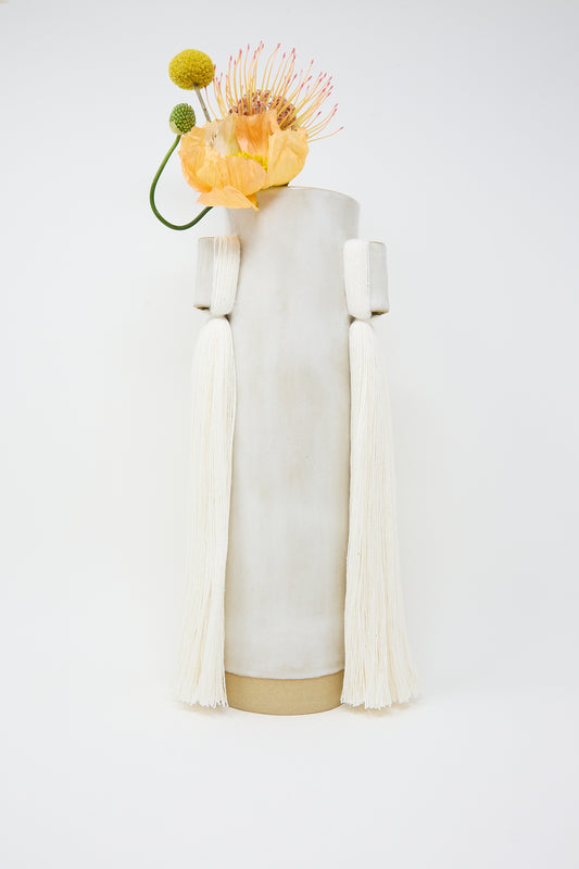 A sculptural piece combining a Karen Tinney handcrafted stoneware Vase #607 in White with draped white tassels and adorned with a whimsical arrangement of flowers and bulbs.