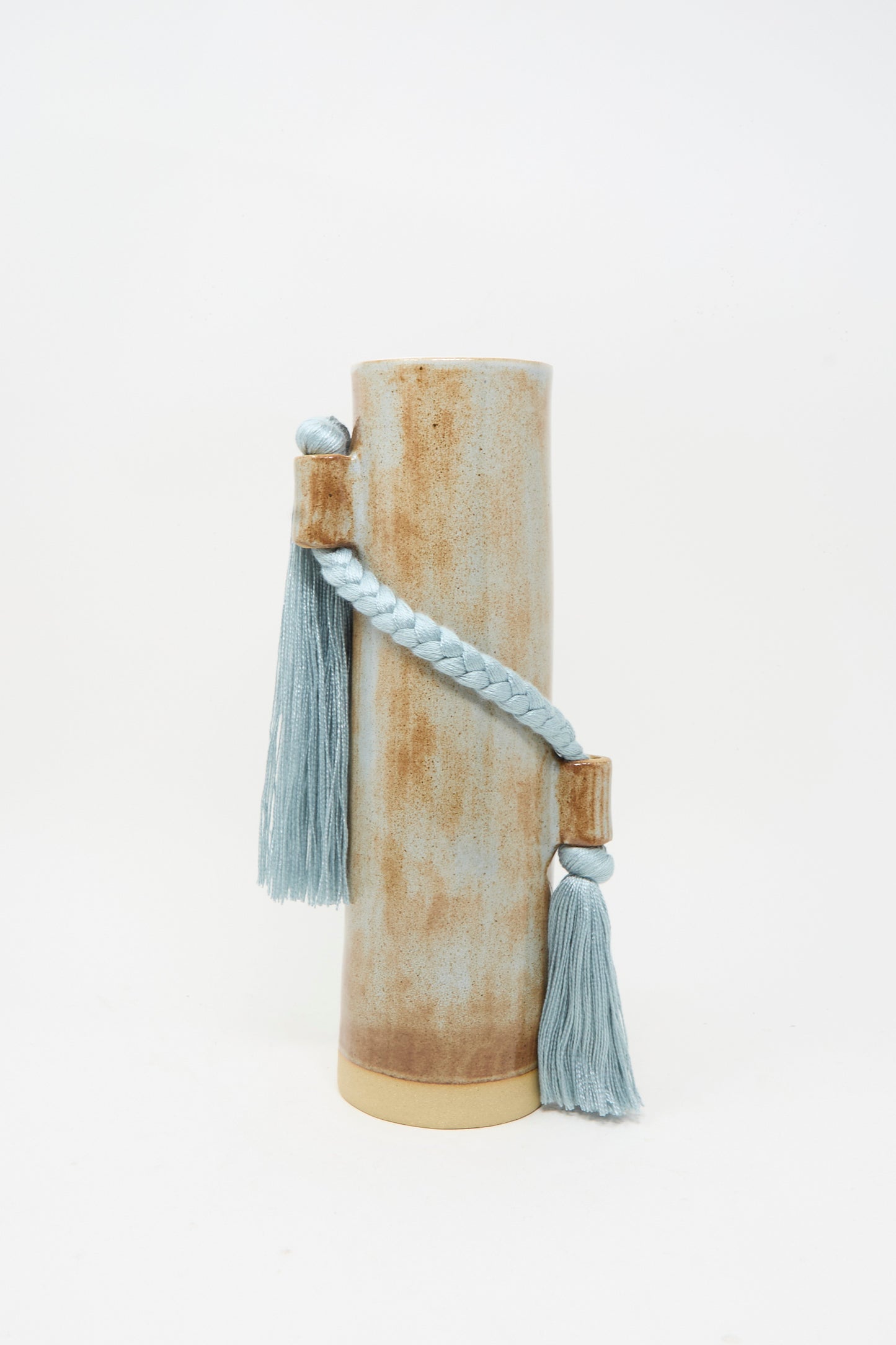 Vase #695 in Blue by Karen Tinney is a tall, cylindrical stoneware vase featuring a brown and beige finish, adorned with a braided blue cord and two blue tassels.