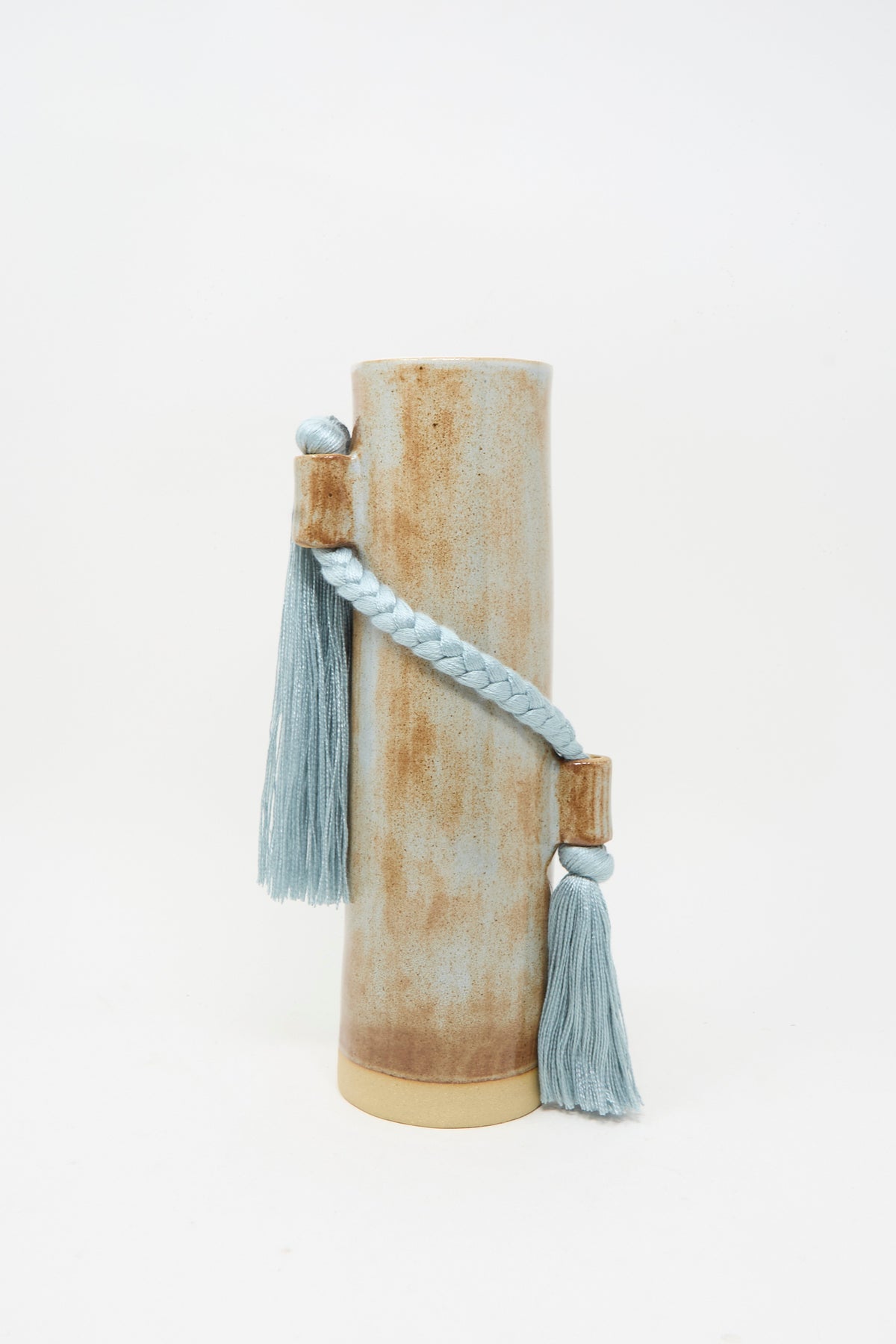 Vase #695 in Blue by Karen Tinney is a tall, cylindrical stoneware vase featuring a brown and beige finish, adorned with a braided blue cord and two blue tassels.