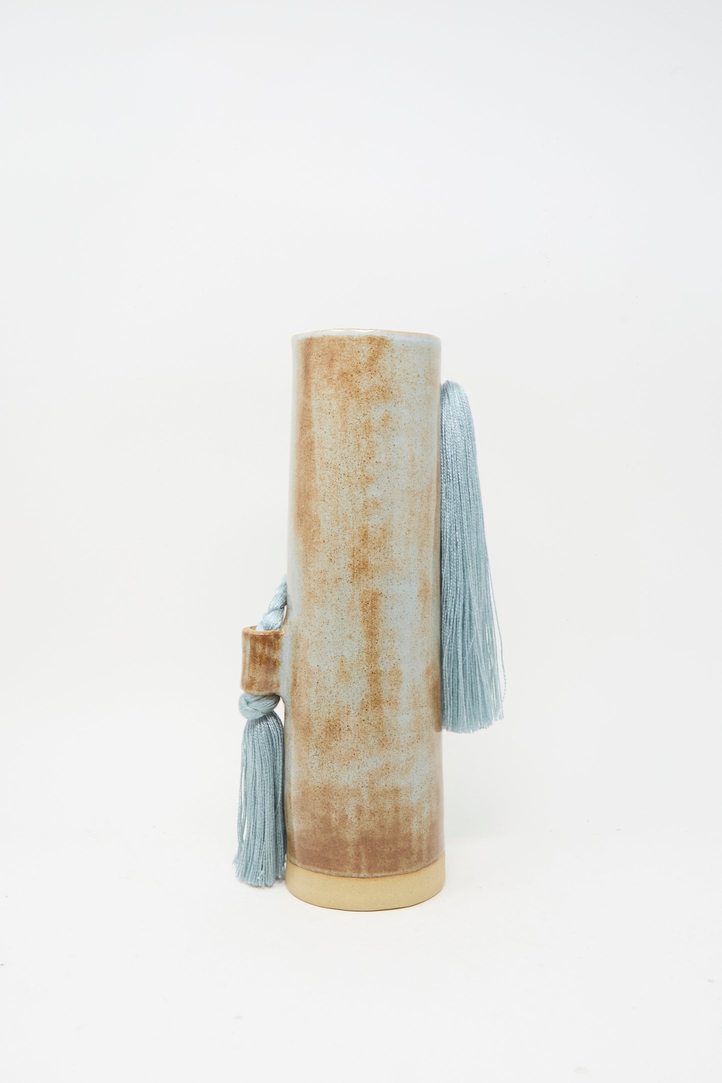 Handcrafted by Karen Tinney, Vase #695 in Blue is a tall, cylindrical stoneware piece with a textured brown surface and two blue tassels attached near the top and side, bringing an artisanal touch to any space.