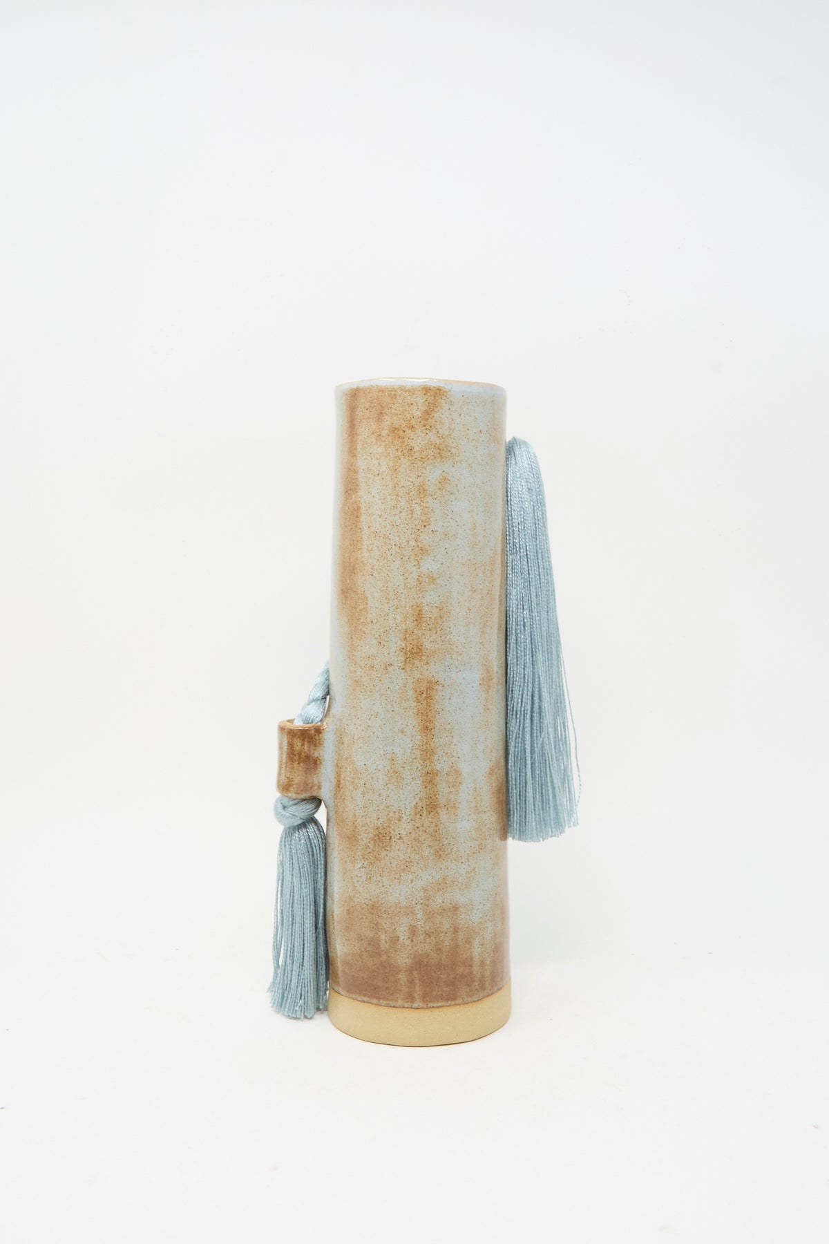 Handcrafted by Karen Tinney, Vase #695 in Blue is a tall, cylindrical stoneware piece with a textured brown surface and two blue tassels attached near the top and side, bringing an artisanal touch to any space.