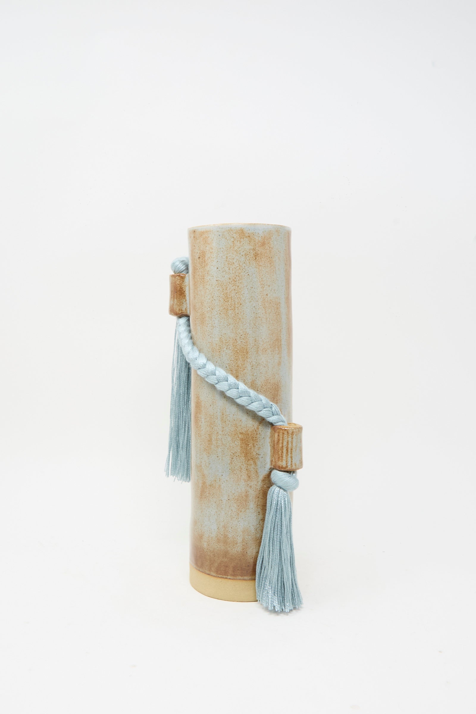 The Karen Tinney Vase #695 in Blue is a cylindrical stoneware piece with a distressed beige finish, adorned with a blue braided cord and two blue tassels attached at the top and bottom.