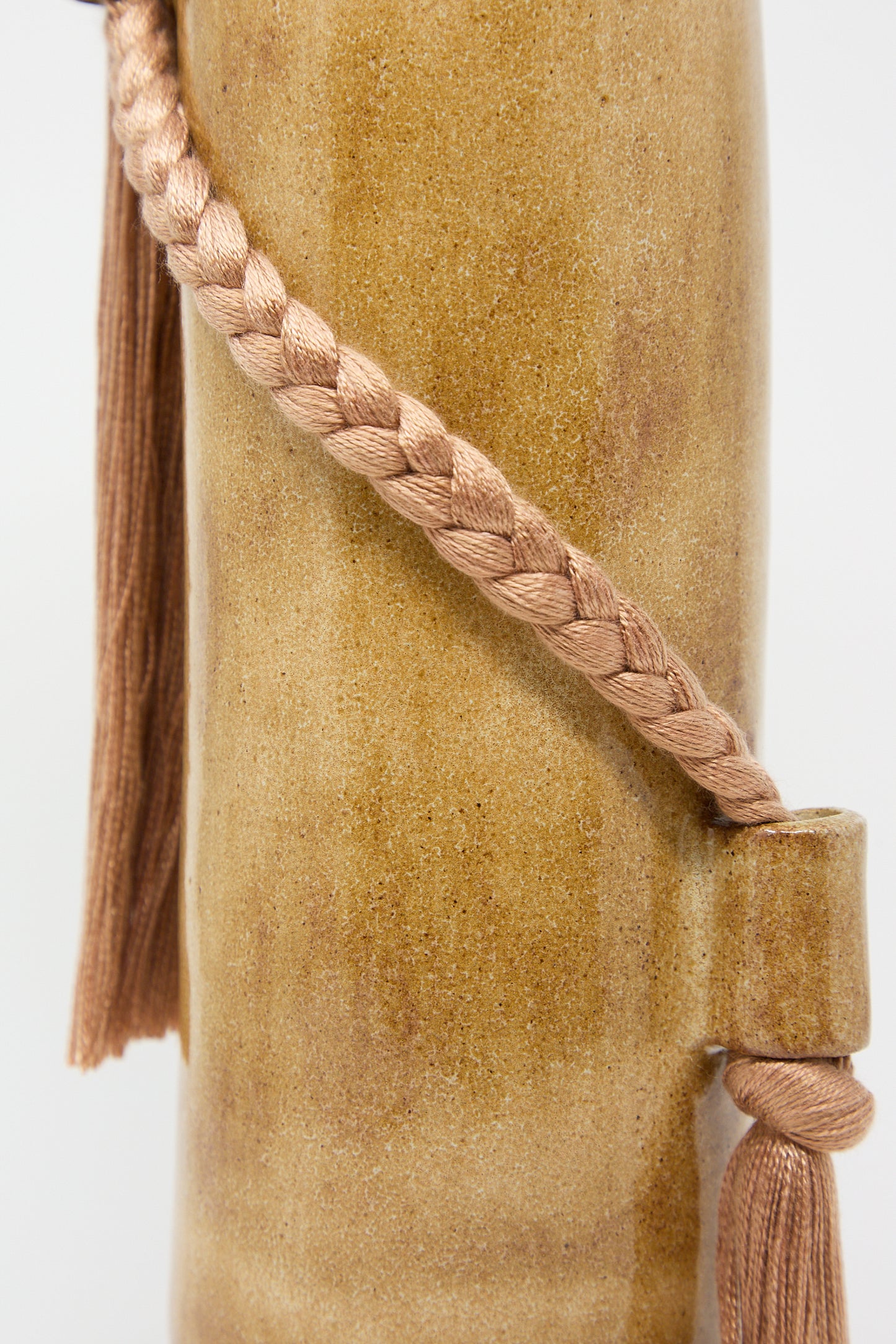 Close-up of a braided tassel on a textured beige surface beside a handcrafted Karen Tinney Vase #695 in Amber.