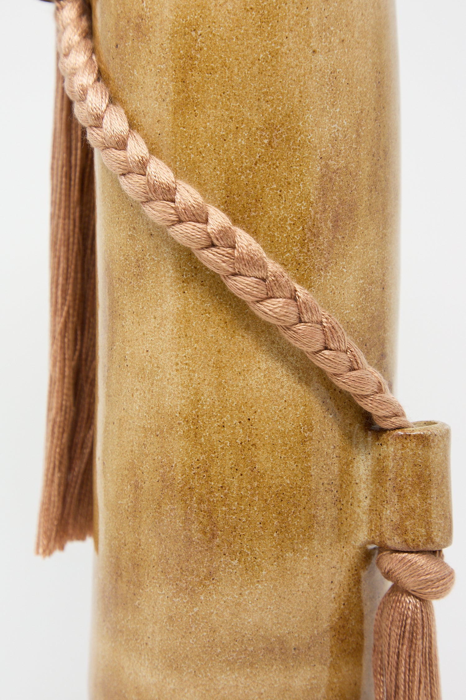 Close-up of a braided tassel on a textured beige surface beside a handcrafted Karen Tinney Vase #695 in Amber.
