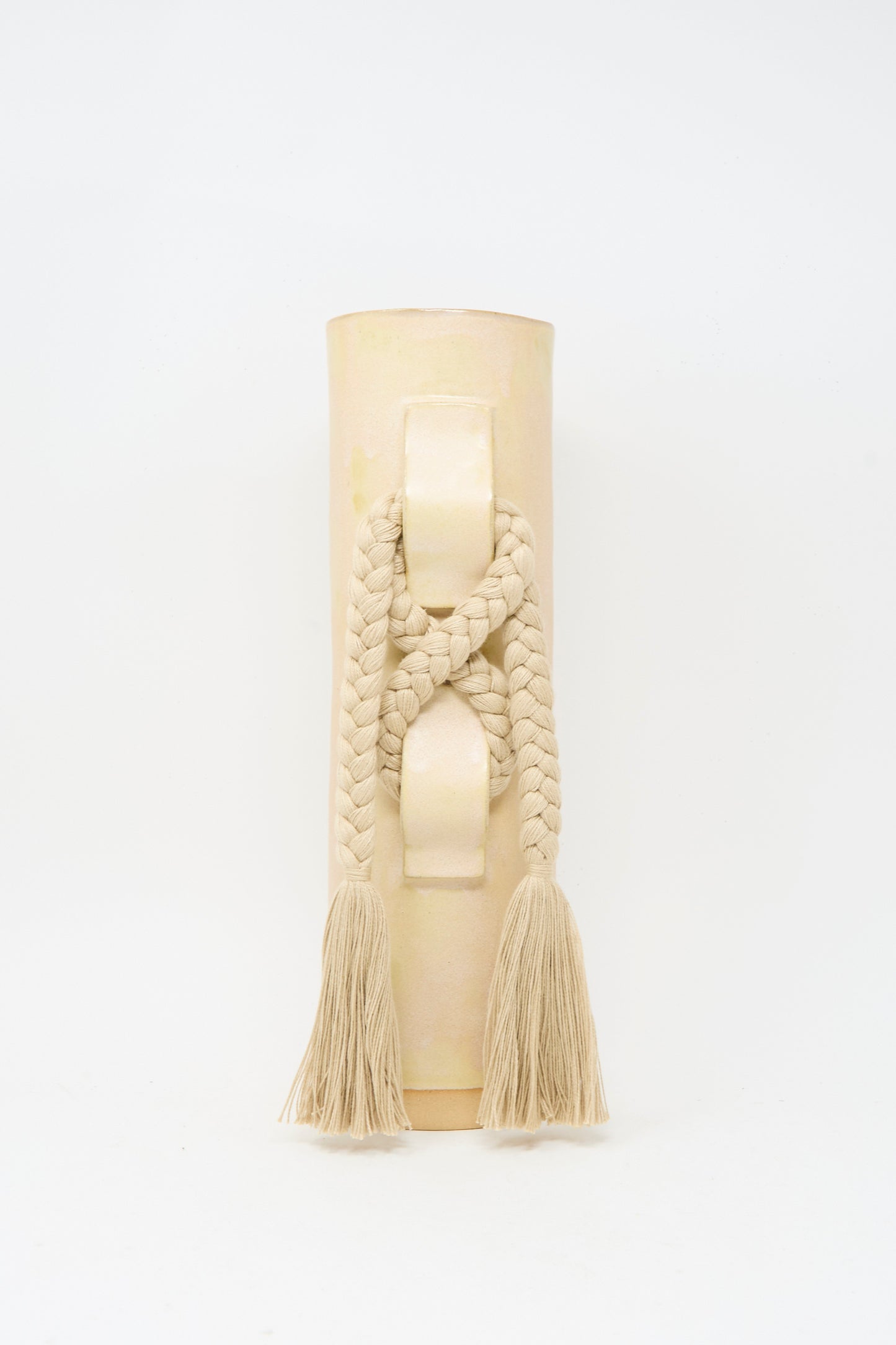 Vase #696 in Tan by Karen Tinney is a beige cylindrical handmade stoneware vase featuring a braided rope and tassels draped over its raised handle, finished in a satin tan glaze and photographed against a plain white background.