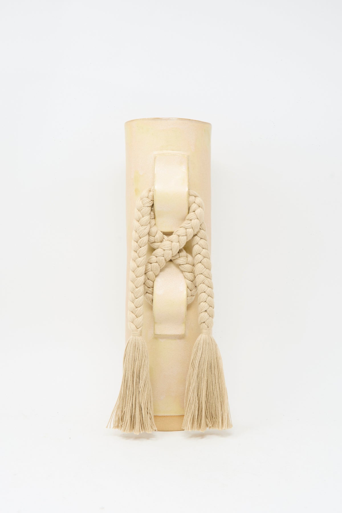 Vase #696 in Tan by Karen Tinney is a beige cylindrical handmade stoneware vase featuring a braided rope and tassels draped over its raised handle, finished in a satin tan glaze and photographed against a plain white background.