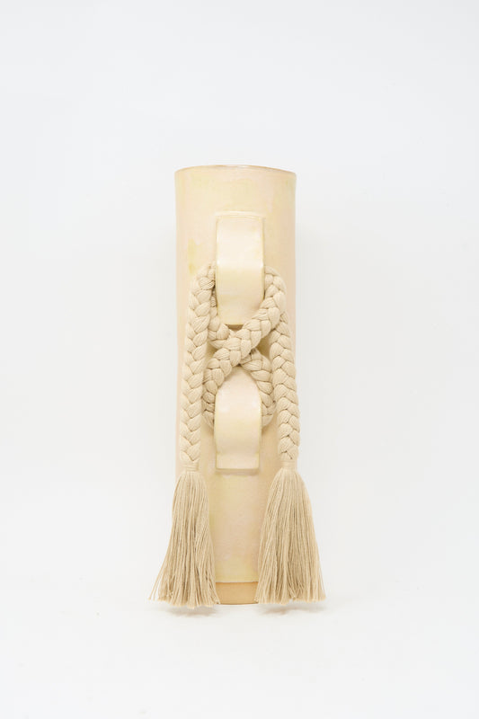 Vase #696 in Tan by Karen Tinney is a beige cylindrical handmade stoneware vase featuring a braided rope and tassels draped over its raised handle, finished in a satin tan glaze and photographed against a plain white background.