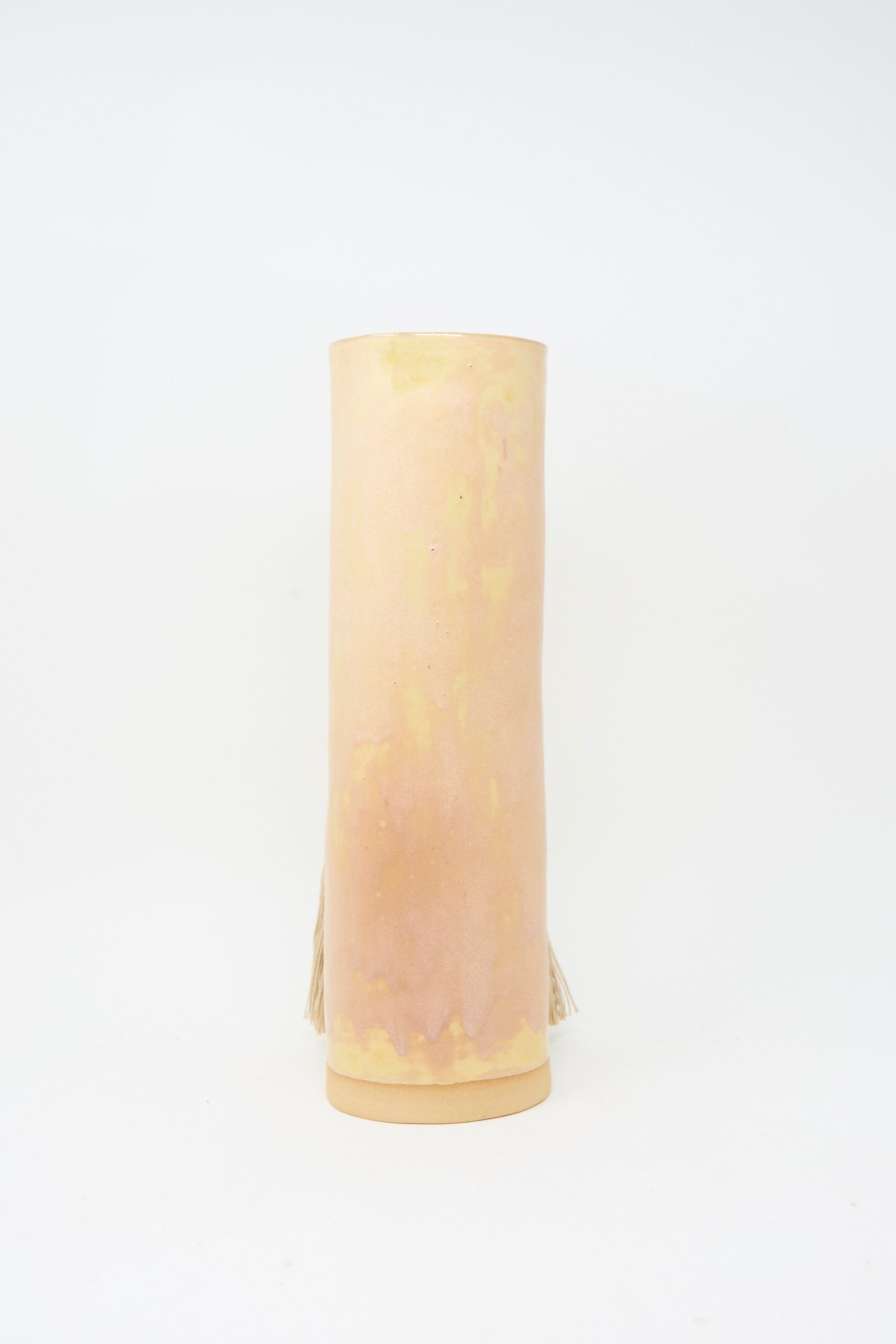 The Karen Tinney Vase #696 in Tan is a tall, cylindrical handmade stoneware vase with a light beige and pinkish hue. It features a smooth surface, subtle fringe detail on the sides, and is finished in a satin tan glaze, set against a plain white background.