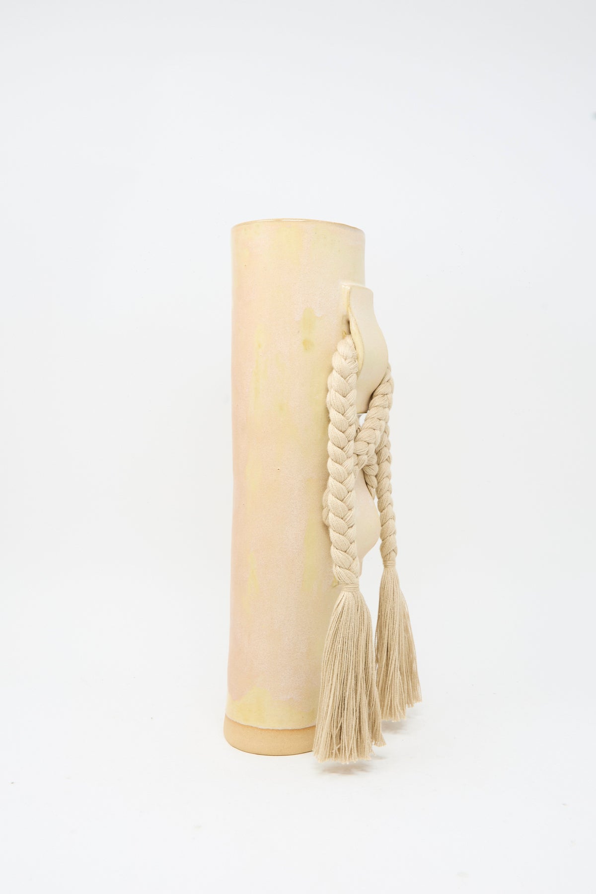 Vase #696 in Tan by Karen Tinney is a tall, cylindrical handmade stoneware vase featuring a muted pastel finish, complemented by a braided rope handle with two tassels and finished with a smooth satin tan glaze, photographed against a plain white background.
