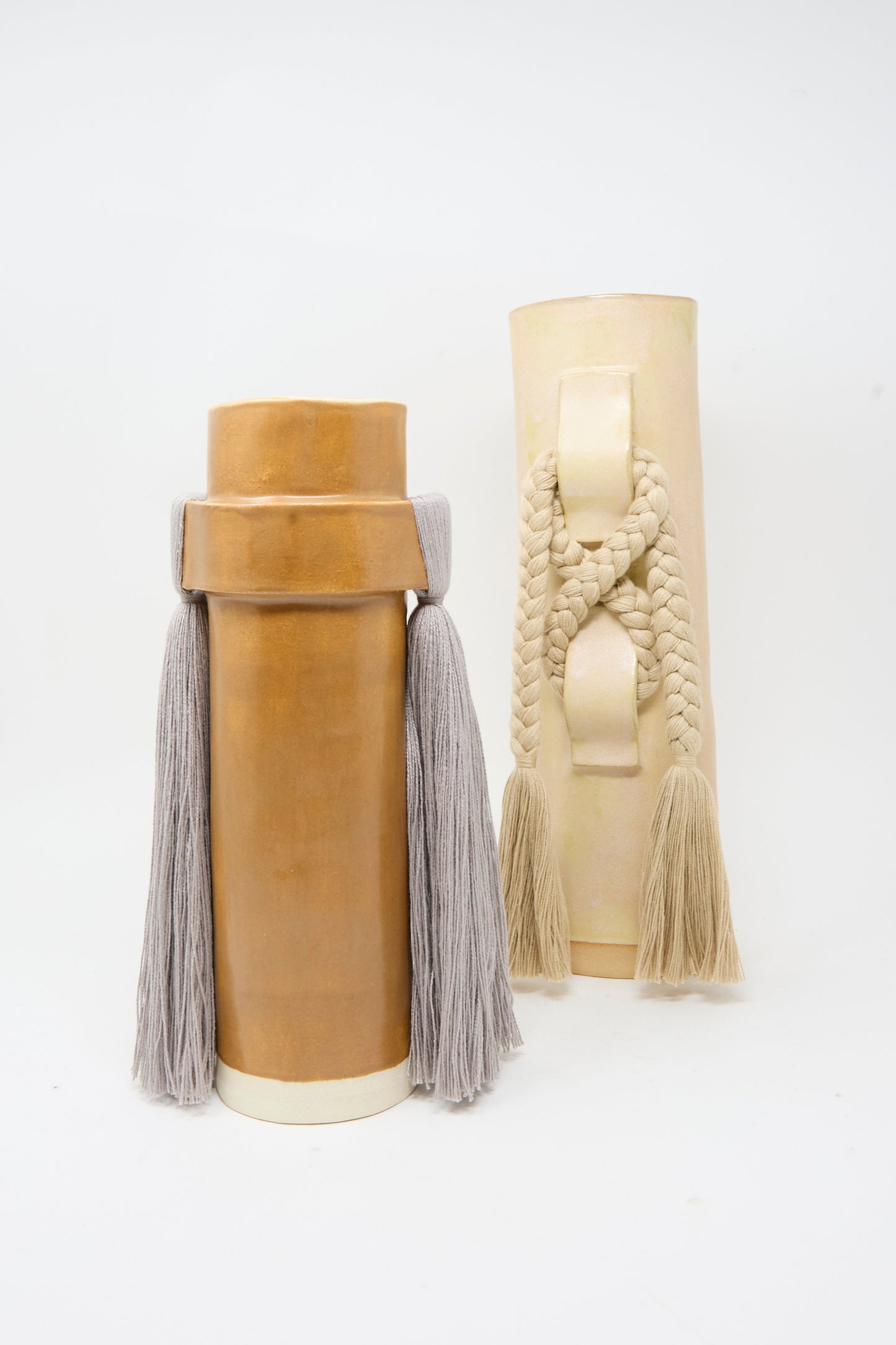 Two cylindrical, handmade stoneware vases with handles by Karen Tinney—Vase #696 in Tan, featuring a satin tan glaze with gray tassels, and a cream variant with braided rope and beige tassels—are artfully displayed on a plain white background. Both pieces are crafted in Vermont.