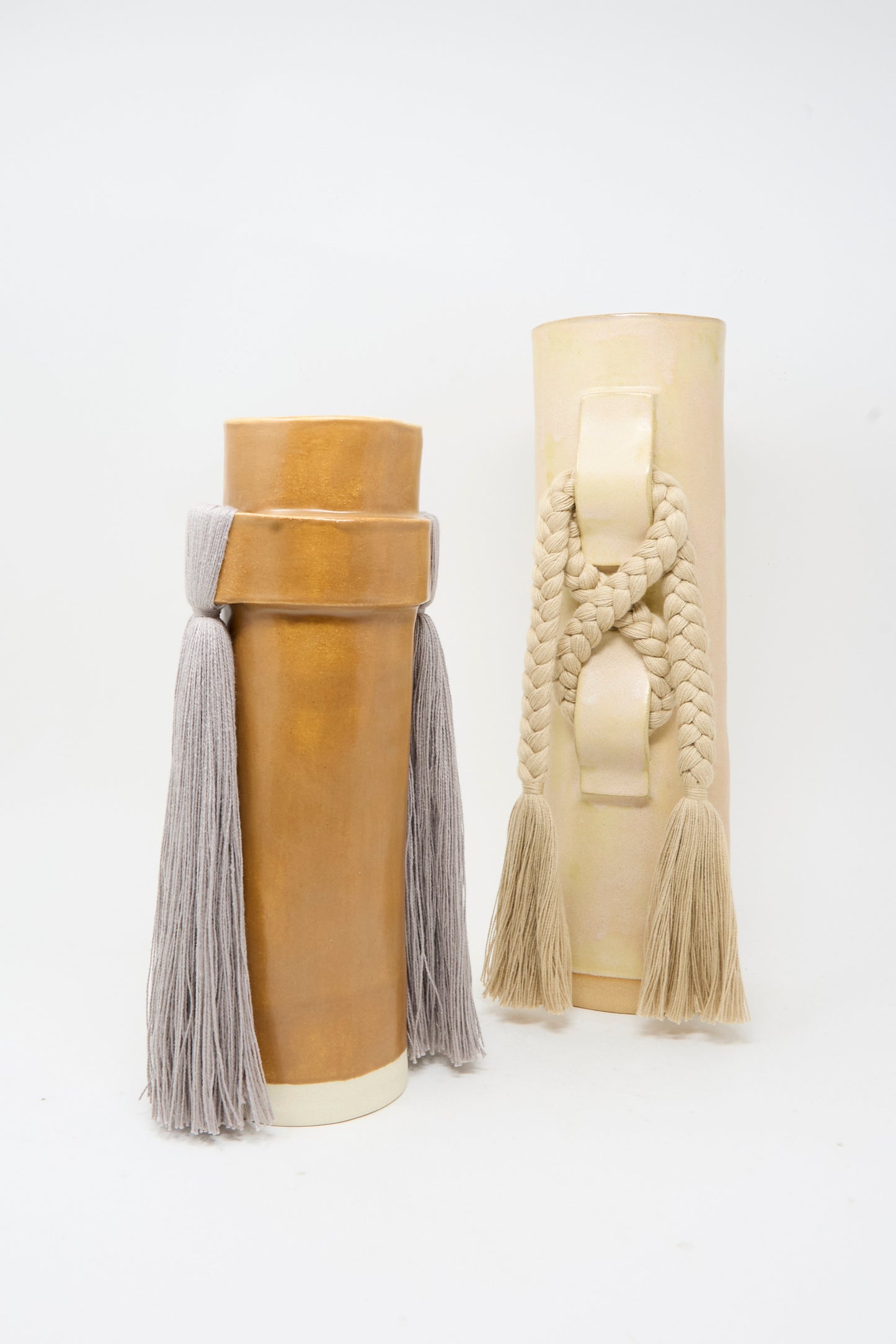 Two tall cylindrical ceramic vases from Karen Tinney, each named Vase #696 in Tan, are adorned with large braided and tasseled rope accents. Set against a plain white background, each handmade stoneware vase features a smooth satin tan glaze and is proudly made in Vermont.