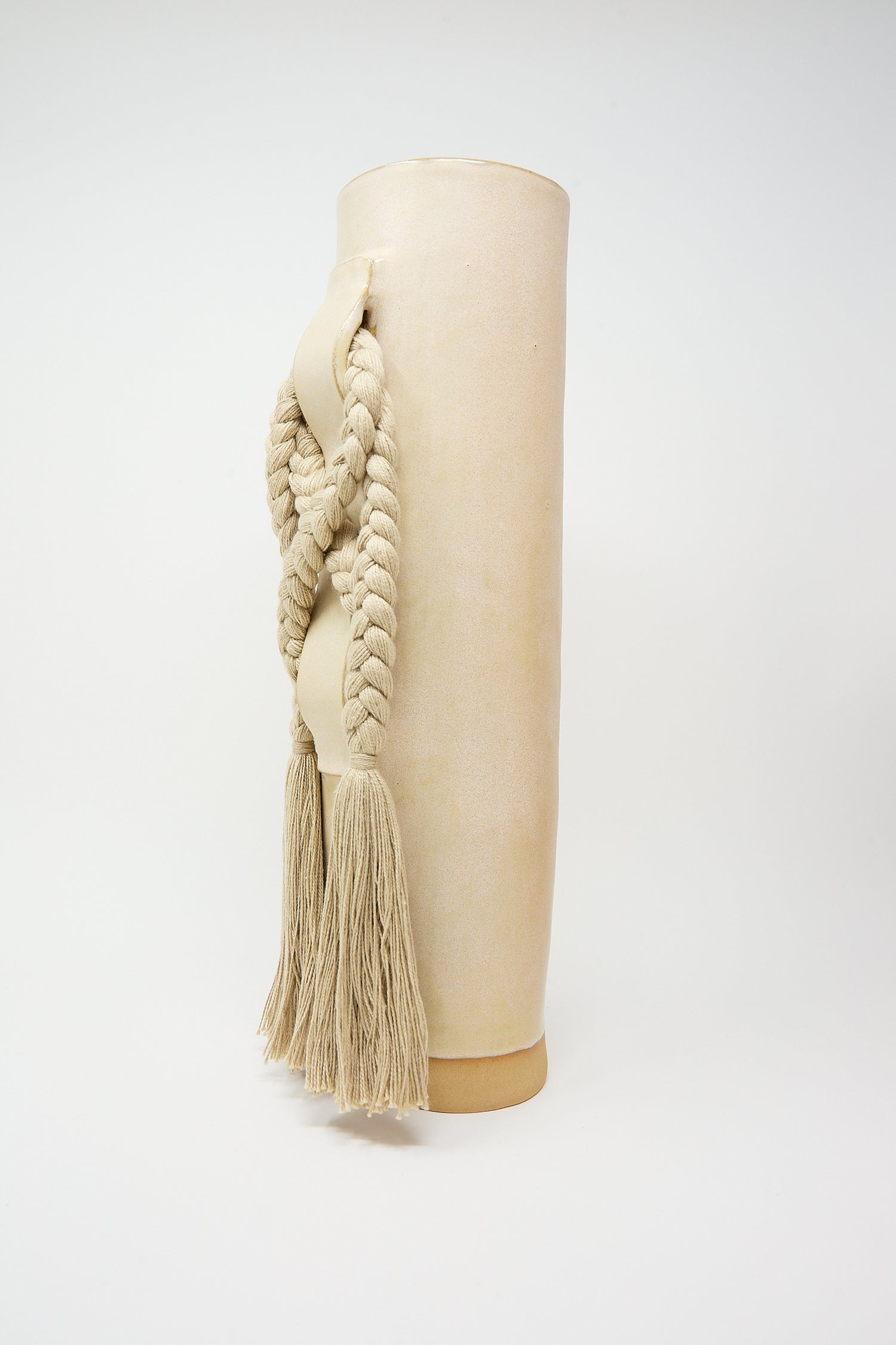 Beige cylindrical handcrafted Vase #696 bolster pillow with braided tassels on a white background by Karen Tinney.