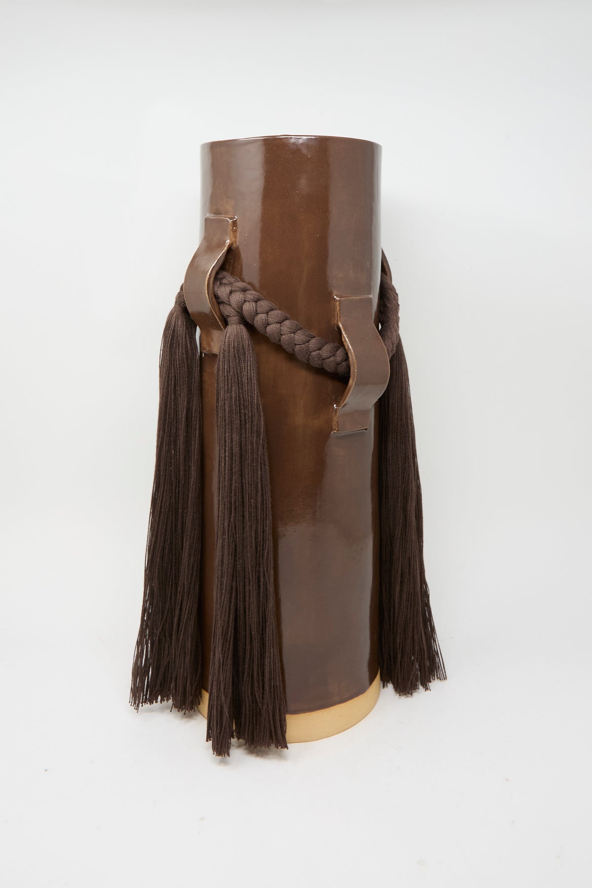 Vase #800 in Brown by Karen Tinney—a tall, cylindrical stoneware vase with a glossy brown glaze, distinctive braided rope detail, and long tassels hanging from each side—stands on a flat surface against a plain white background, showcasing exceptional handmade craftsmanship.