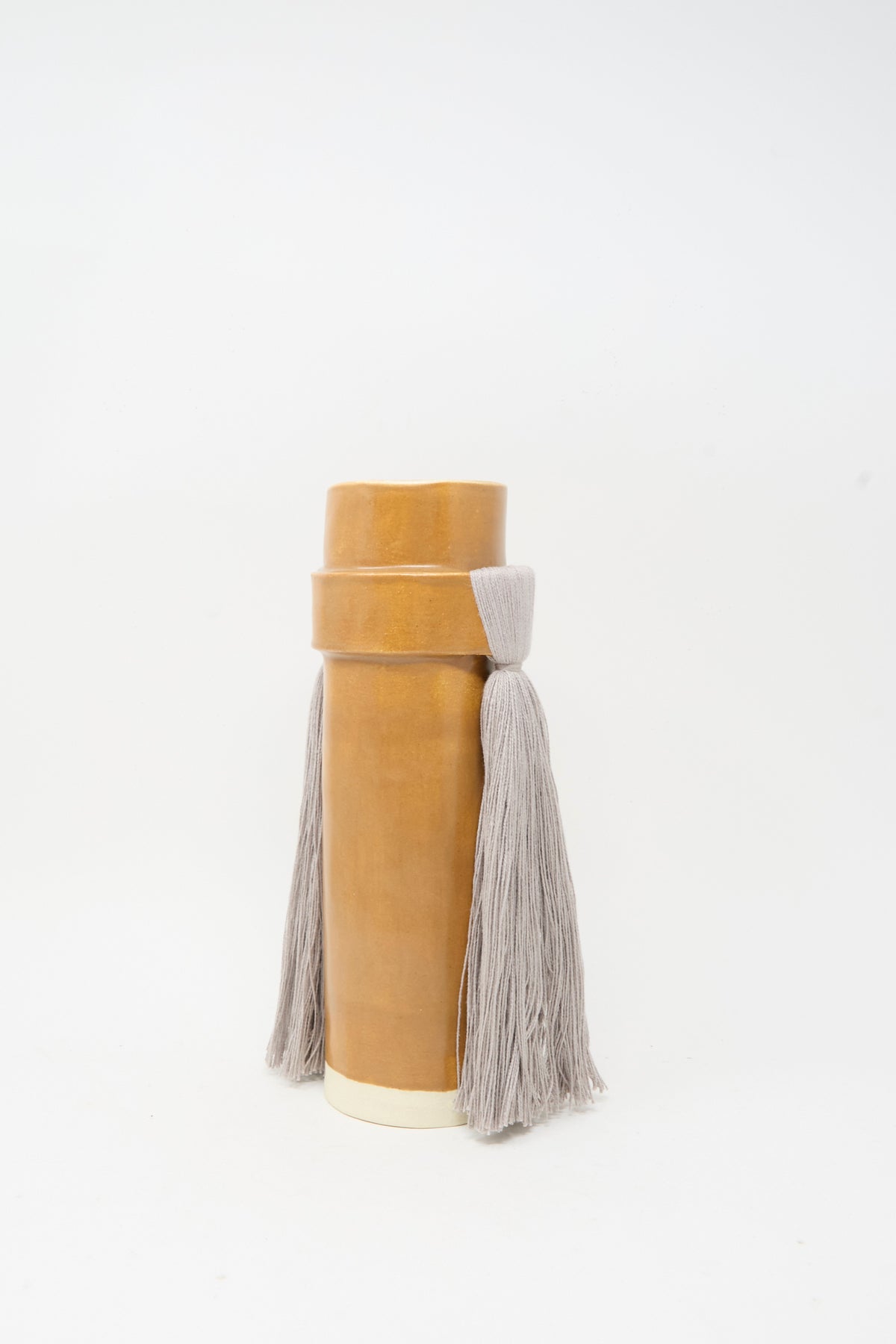 The Karen Tinney Vase #825 in Brown and Gray is a cylindrical tan vessel with a white base, finished in a satin brown glaze. It features a lid and is adorned with two long, thick gray tassels hanging from the sides, embodying the look of a handmade decorative piece.