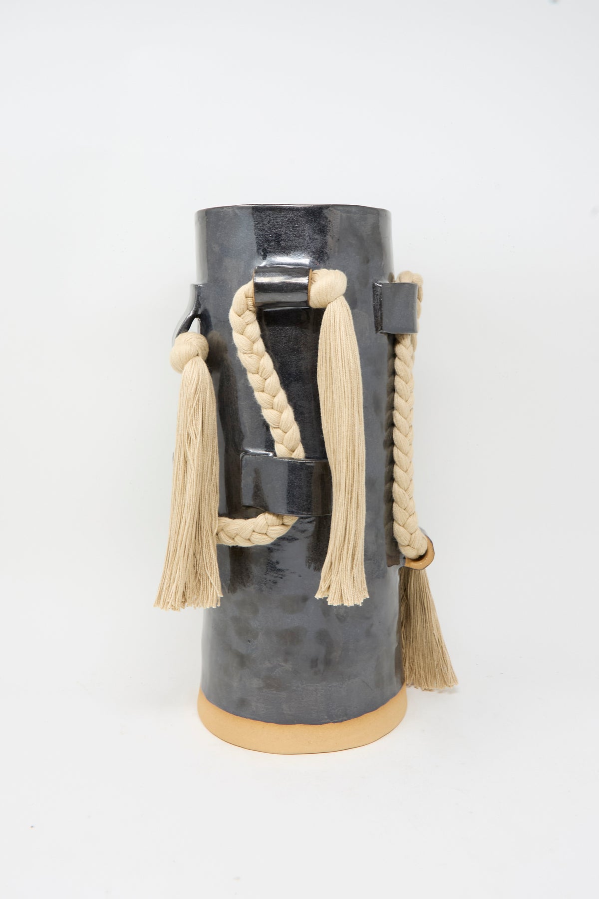 Vase #846 in Black and Tan by Karen Tinney is a tall, cylindrical stoneware vase featuring a glossy black glaze, adorned with braided and tasseled beige ropes that wrap around and hang from its sides, shown against a plain white background.