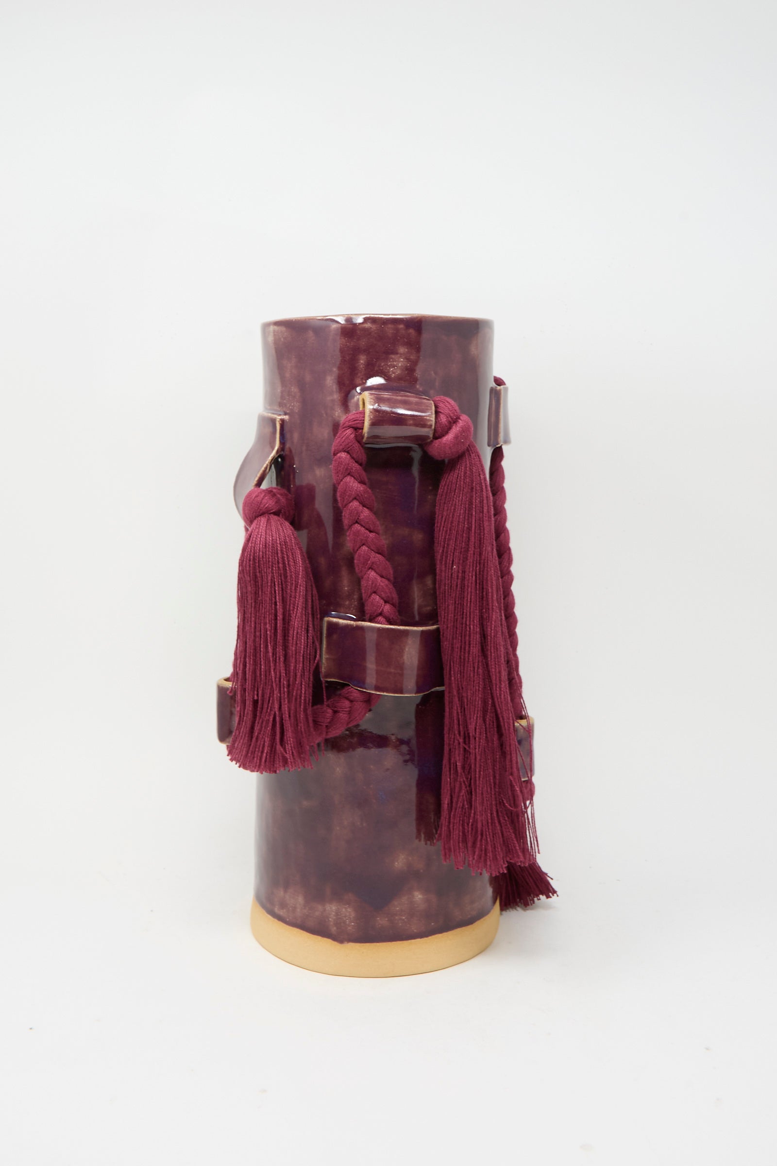 Vase #846 in Plum by Karen Tinney is a tall, glossy maroon stoneware vase with an attached braided cord and tassels, featuring a beige base and striking burgundy glaze, photographed against a plain white background.