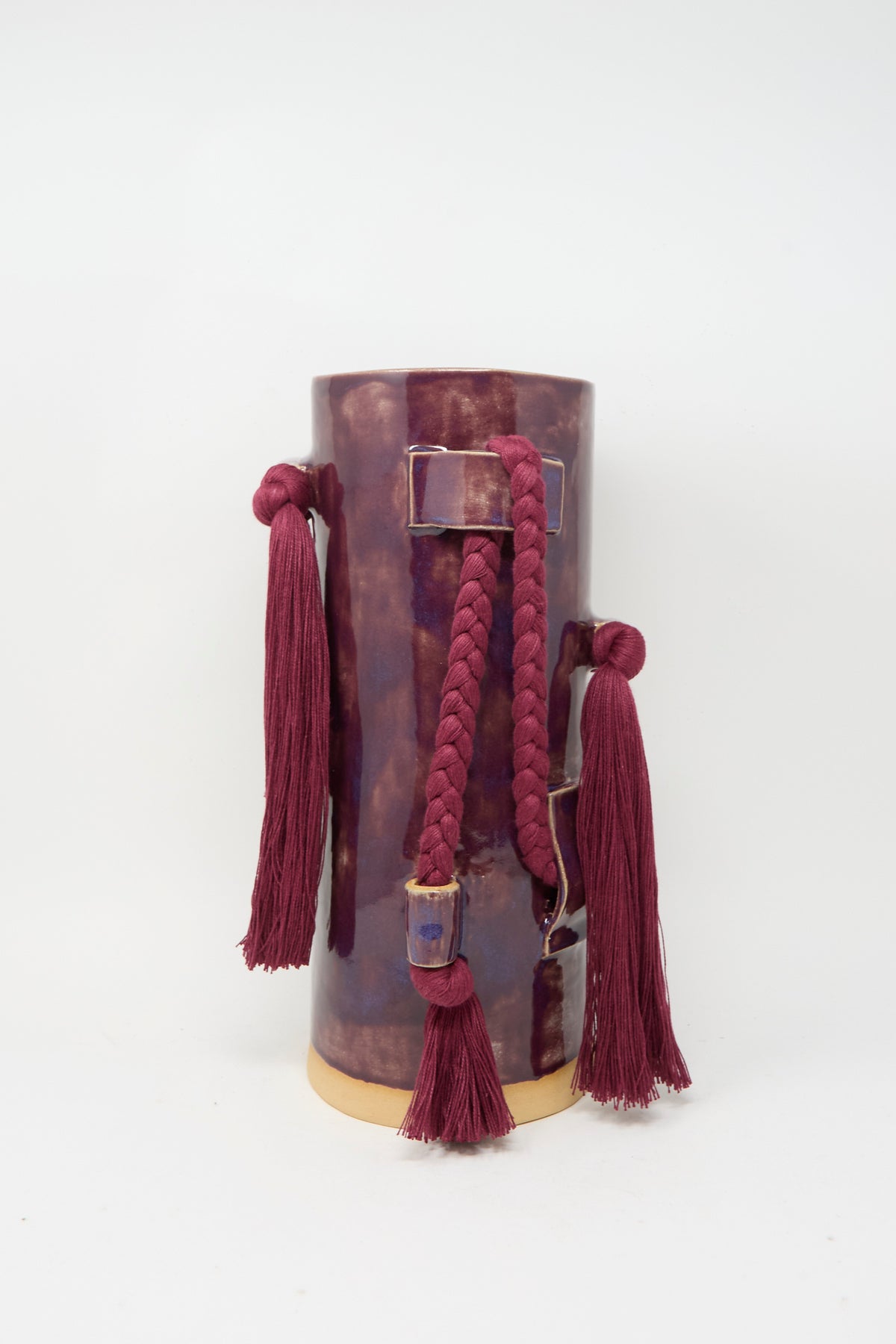 The Karen Tinney Vase #846 in Plum is a handmade stoneware vase with a glossy plum finish, adorned with matching tassels and a braided cord, set off by a contrasting beige base.
