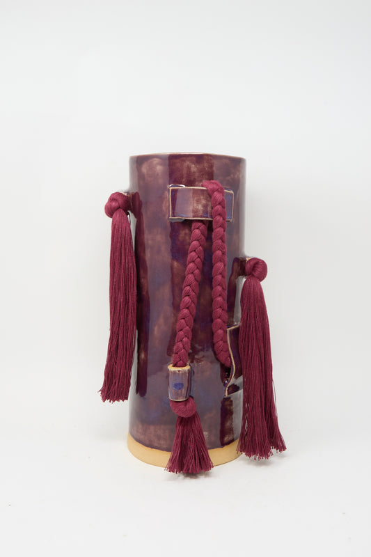 The Karen Tinney Vase #846 in Plum is a handmade stoneware vase with a glossy plum finish, adorned with matching tassels and a braided cord, set off by a contrasting beige base.