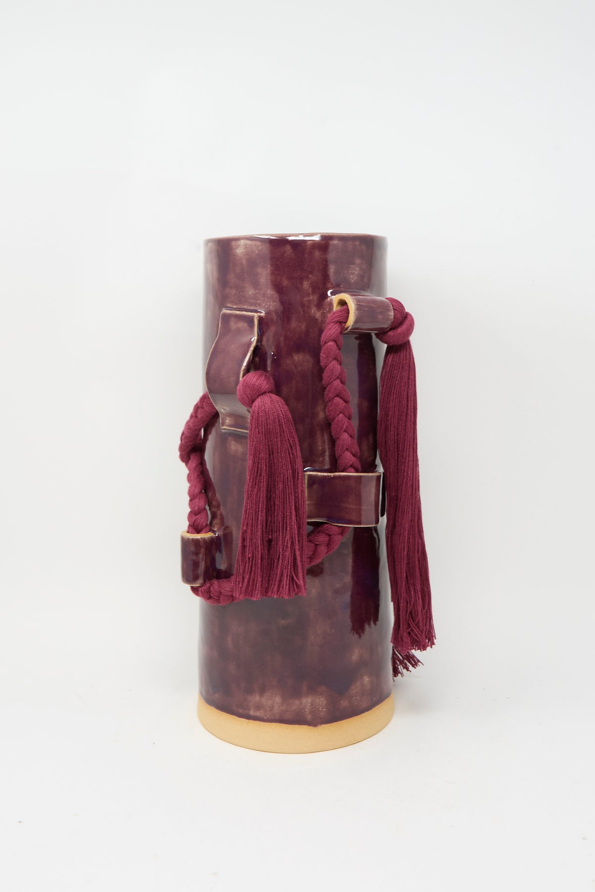 Karen Tinney’s Vase #846 in Plum, a tall cylindrical stoneware piece with braided cord, tassels, and leather strap accents, stands on a light background.
