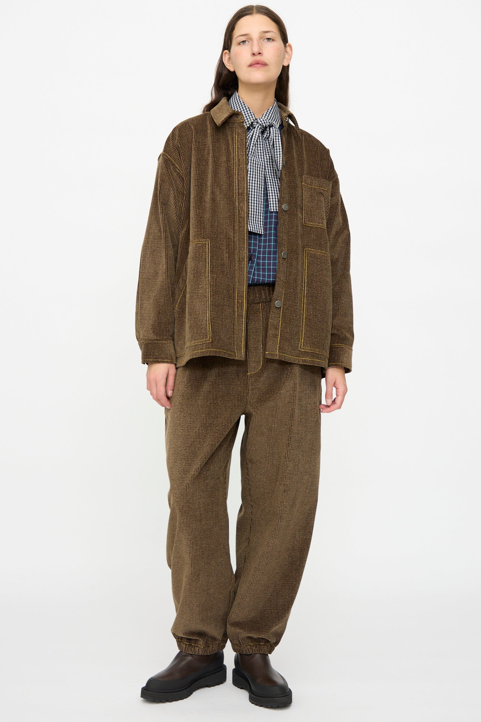 A person stands against a white background wearing the KasMaria Corduroy Oversized Shirt Jacket in Brown, paired with matching pants, a checkered shirt, and black shoes.