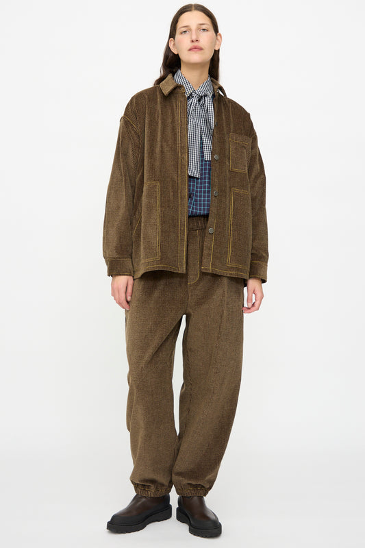 A person stands against a white background wearing the KasMaria Corduroy Oversized Shirt Jacket in Brown, paired with matching pants, a checkered shirt, and black shoes.