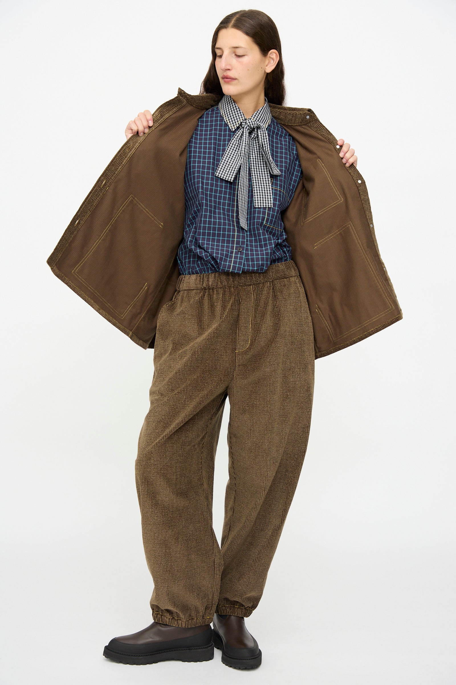 A woman stands with eyes closed, wearing the KasMaria Corduroy Oversized Shirt Jacket in Brown over a blue plaid shirt with a large necktie, paired with loose brown corduroy pants and black shoes.