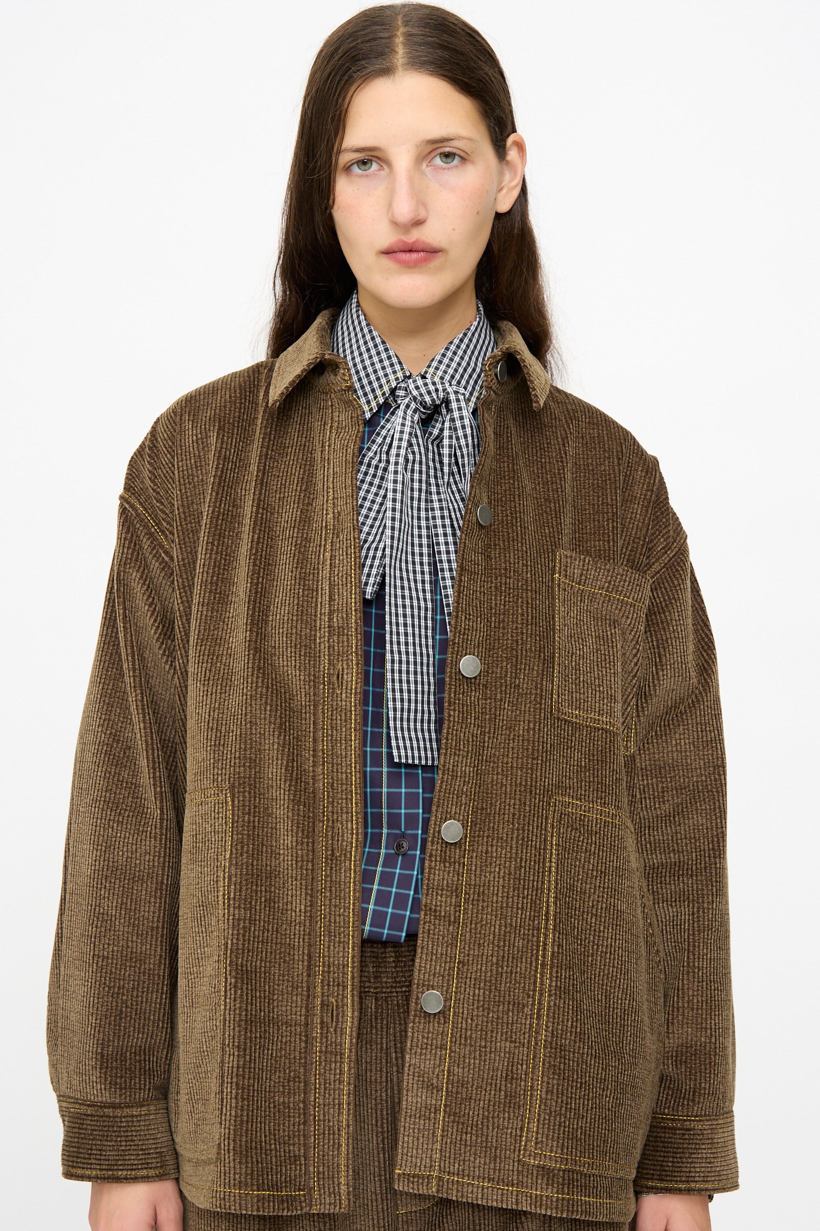 A person with long brown hair wears the KasMaria Corduroy Oversized Shirt Jacket in Brown over a checked shirt with a bow at the collar, standing against a plain white background.