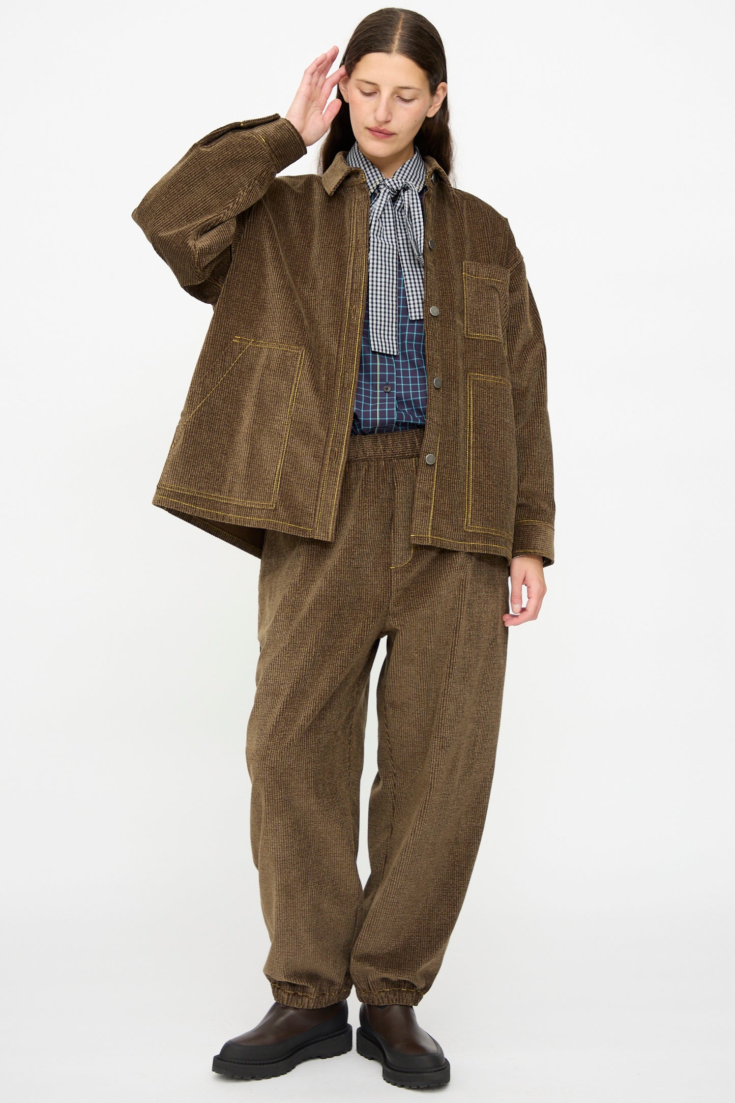 A person wearing the KasMaria Corduroy Oversized Shirt Jacket in Brown, paired with matching pants, an oversized Japanese cotton plaid shirt, and a black-and-white checkered scarf, stands against a plain white background.
