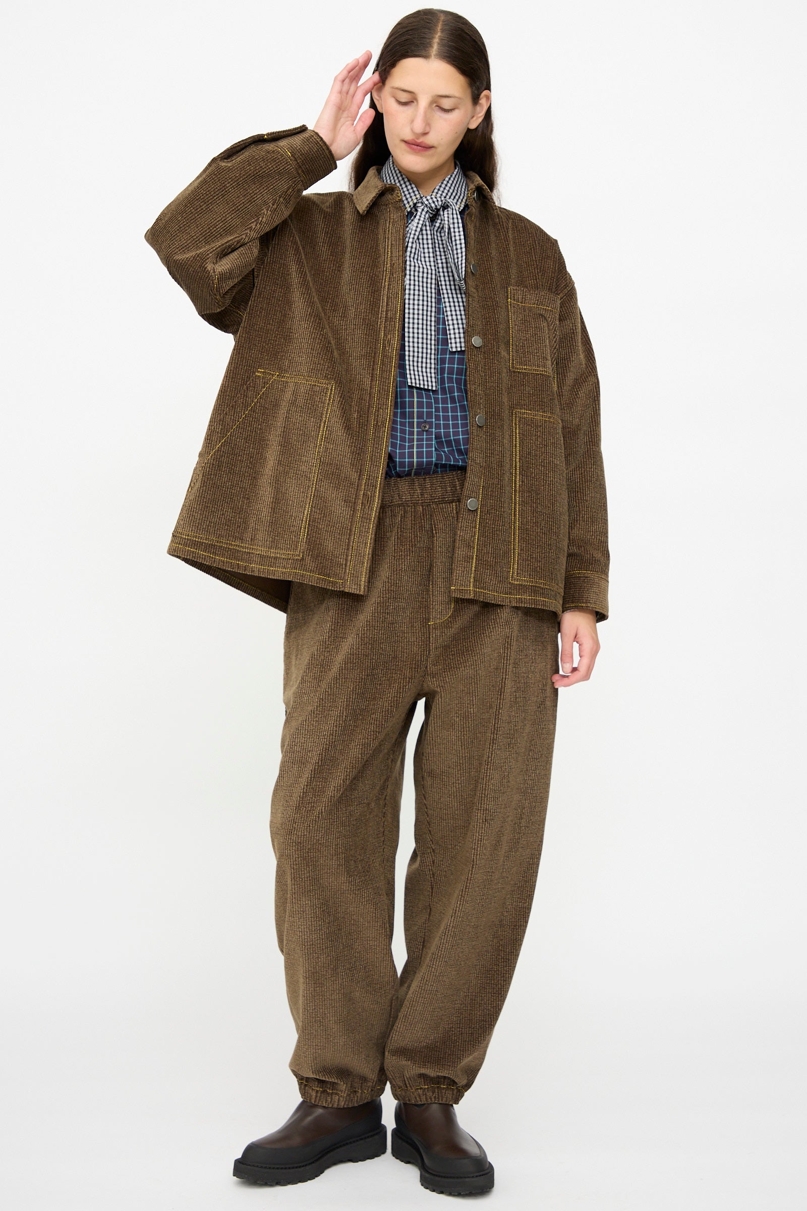 A person wearing the KasMaria Corduroy Oversized Shirt Jacket in Brown, paired with matching pants, an oversized Japanese cotton plaid shirt, and a black-and-white checkered scarf, stands against a plain white background.