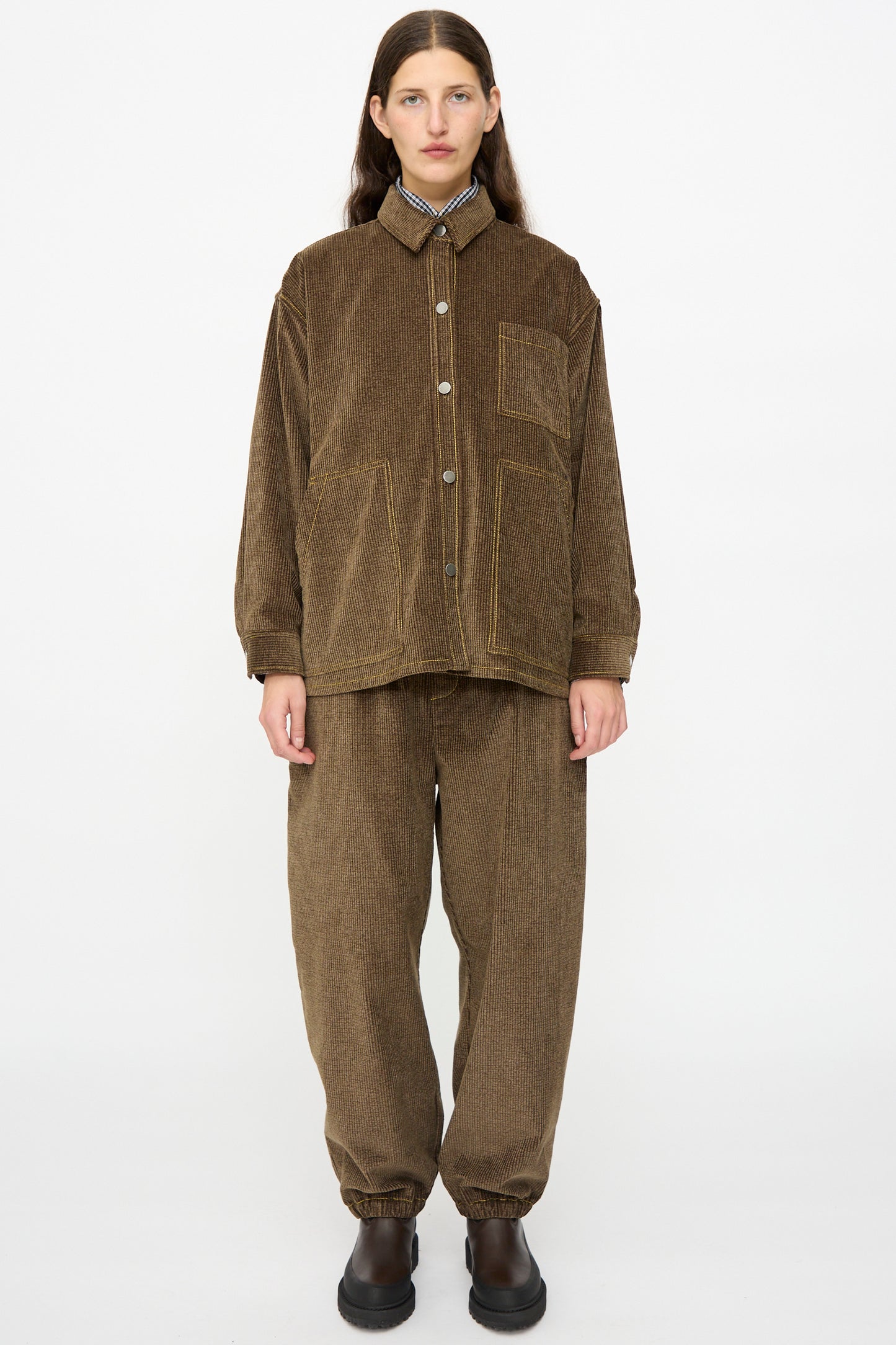 A person faces forward wearing the KasMaria Corduroy Oversized Shirt Jacket in Brown with matching loose pants and black slip-on shoes, standing against a plain white background.