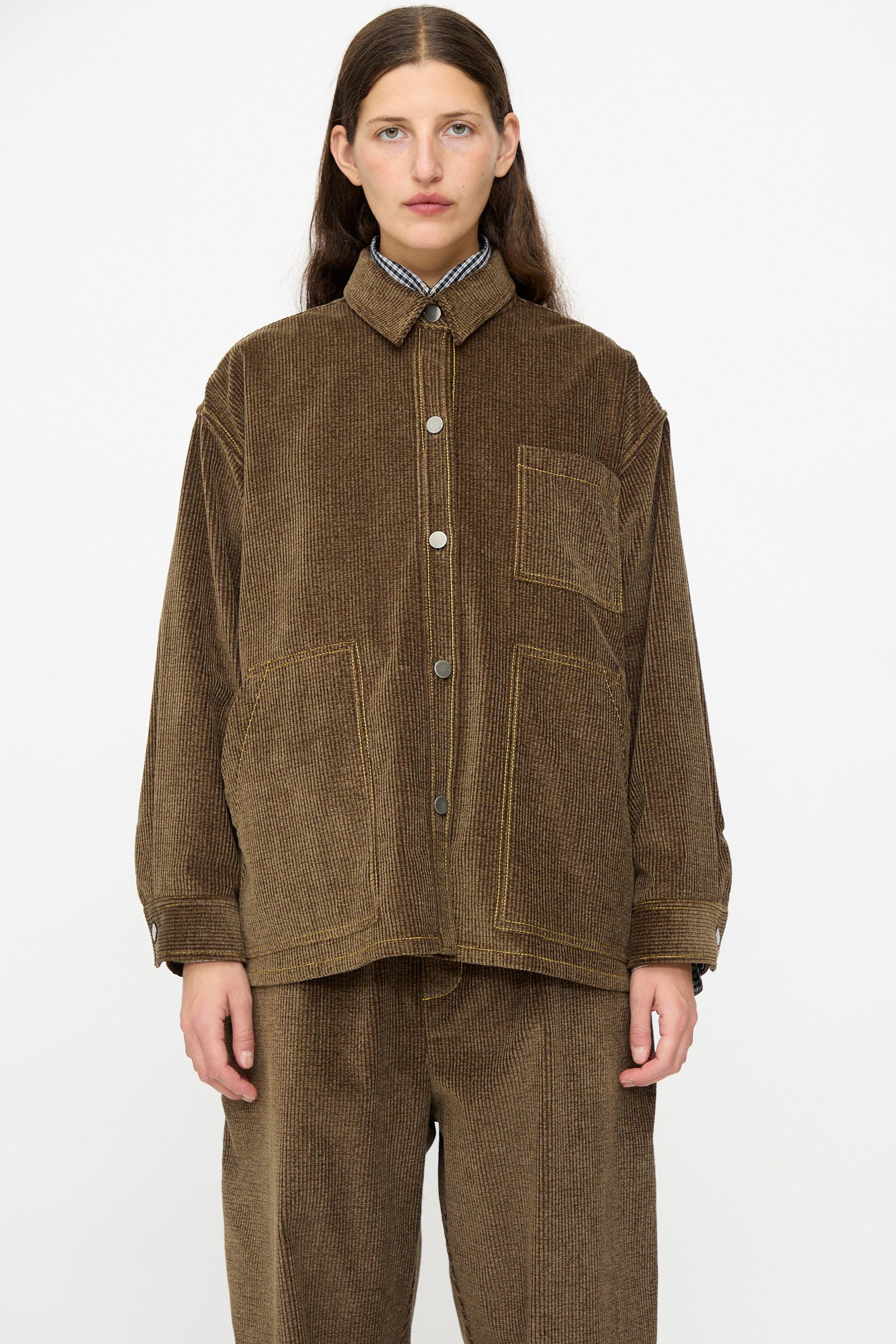 A person stands against a plain background wearing the KasMaria Corduroy Oversized Shirt Jacket in Brown, featuring large front pockets and matching pants, all crafted from soft Japanese cotton.