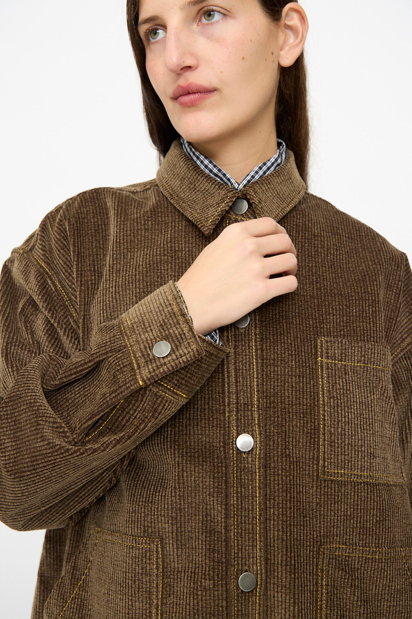 A person wearing the Corduroy Oversized Shirt Jacket in Brown by KasMaria with metal buttons and yellow stitching, holding the collar and looking slightly to the side.