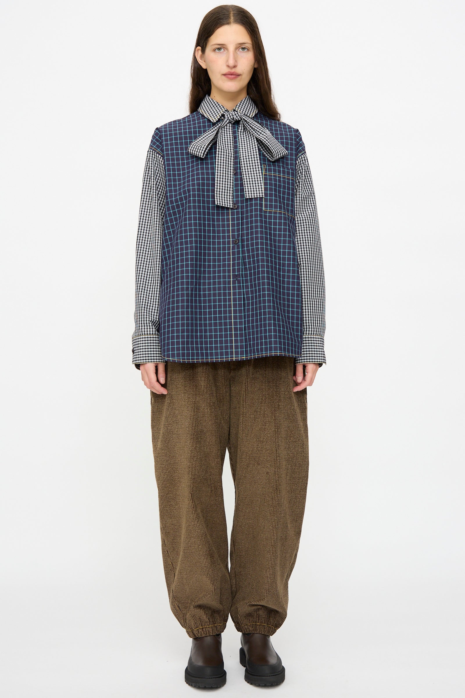 A person stands against a white background wearing KasMaria's Corduroy Sweatpant in Brown, paired with a blue plaid vest over a checkered shirt, a bow tie, and black shoes.