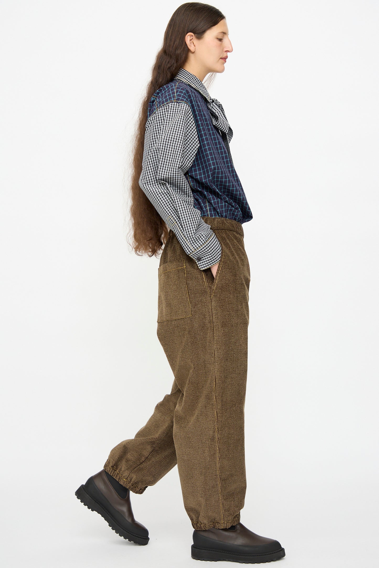 A person with long hair walks in profile, wearing a checkered shirt, blue plaid vest, KasMaria Corduroy Sweatpant in Brown made from Japanese cotton, and black boots against a plain white background.