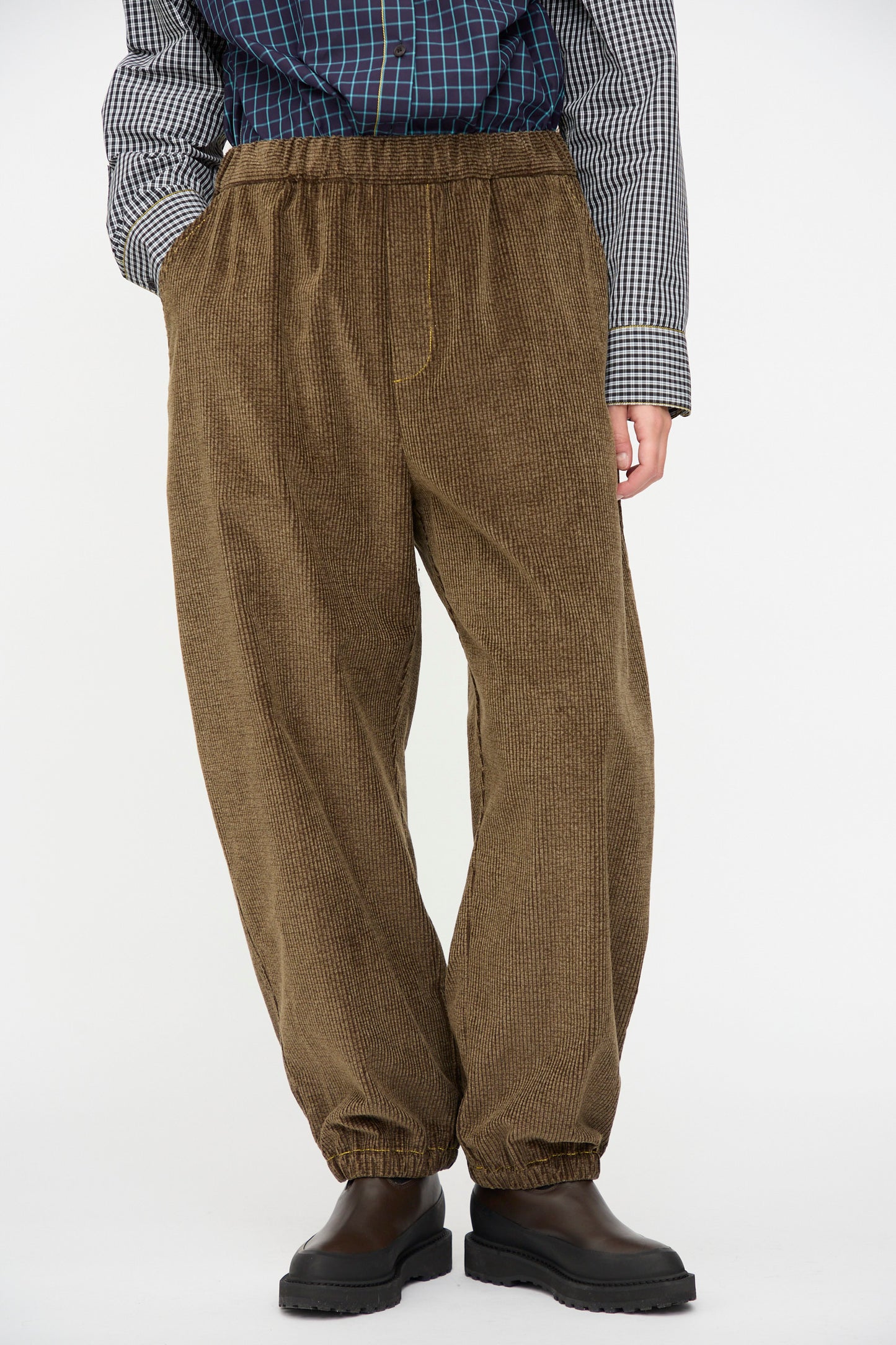 A person stands against a plain background wearing KasMaria's Corduroy Sweatpant in Brown, paired with a checkered Japanese cotton shirt and black boots.