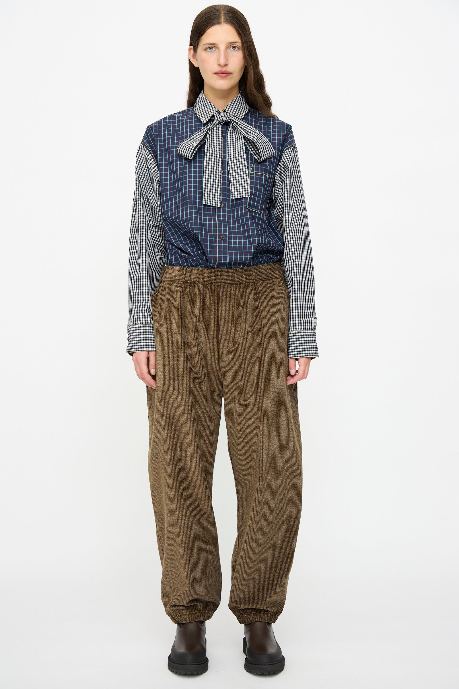 A woman faces forward against a white background wearing KasMaria's Corduroy Sweatpant in Brown, paired with a plaid blouse with a large bow and black shoes.