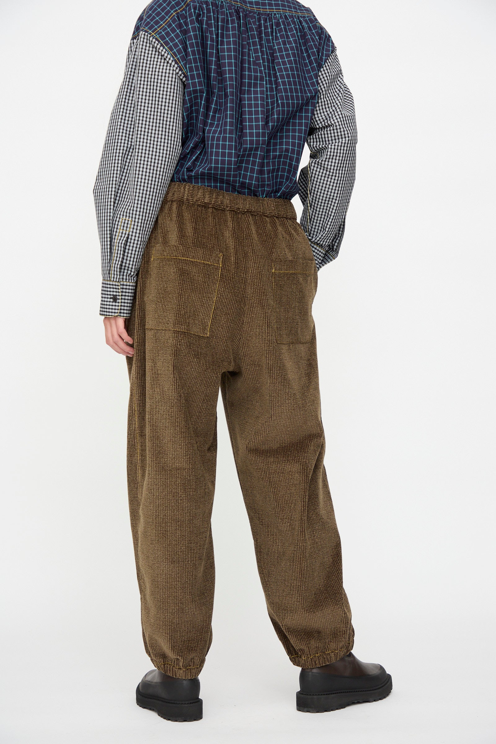 A person stands facing away in KasMaria’s Corduroy Sweatpant in Brown—relaxed fit, Japanese cotton, back pockets—paired with a blue plaid shirt and black shoes, against a plain white background.
