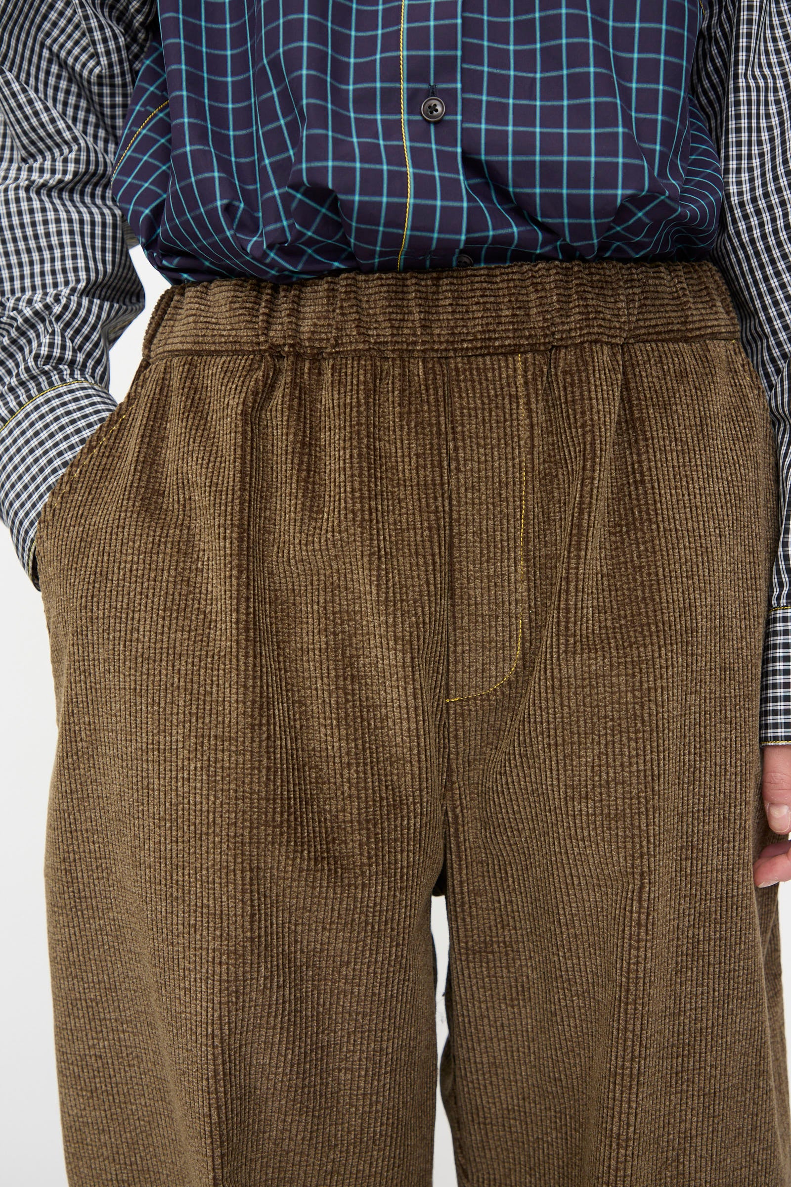 A person wears the KasMaria Corduroy Sweatpant in Brown, featuring a relaxed fit and elastic waistband, paired with a blue, black, and white checkered Japanese cotton button-up shirt.